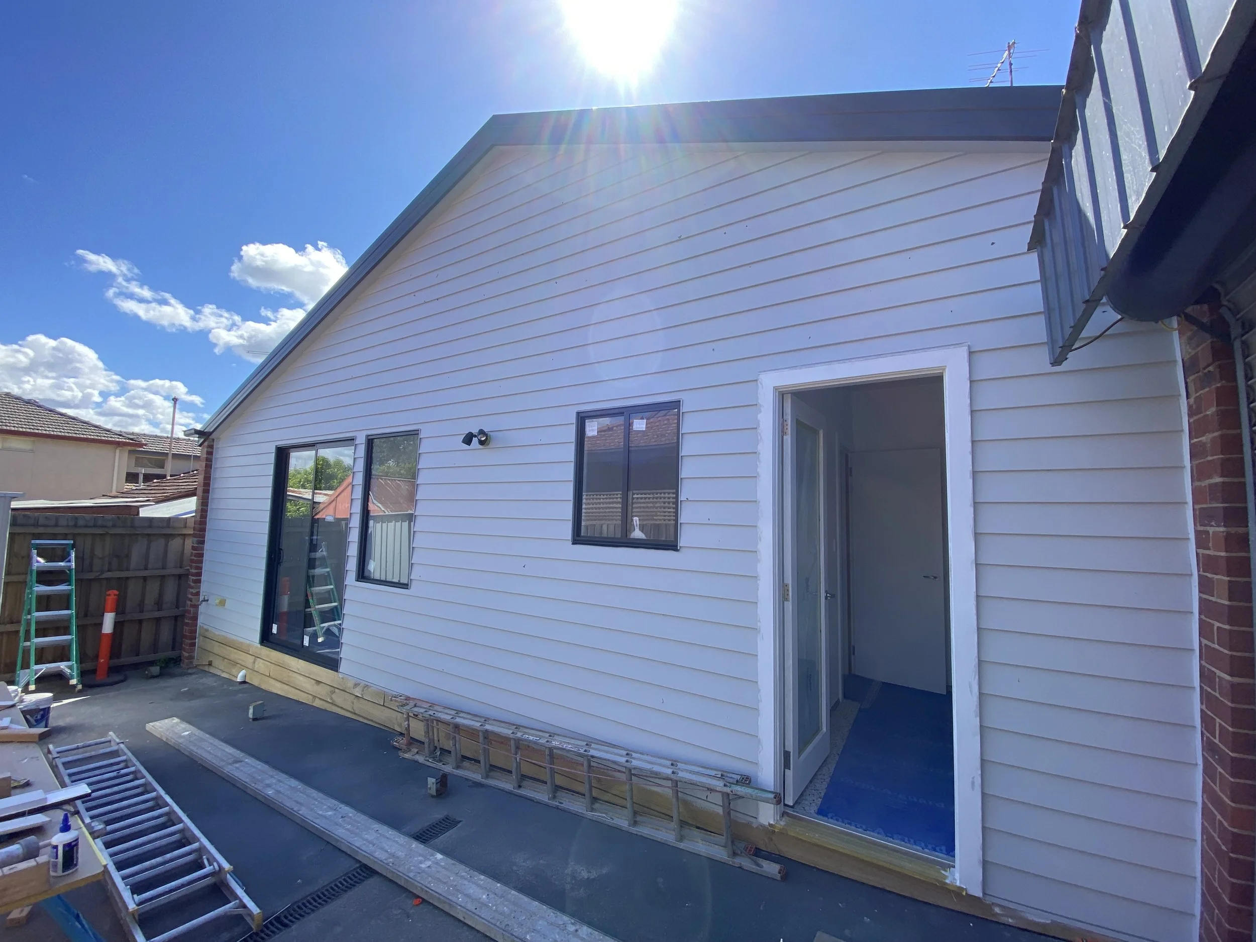 Backyard of a house under construction with vinyl siding, an open sliding door, and construction tools and materials on the ground.
