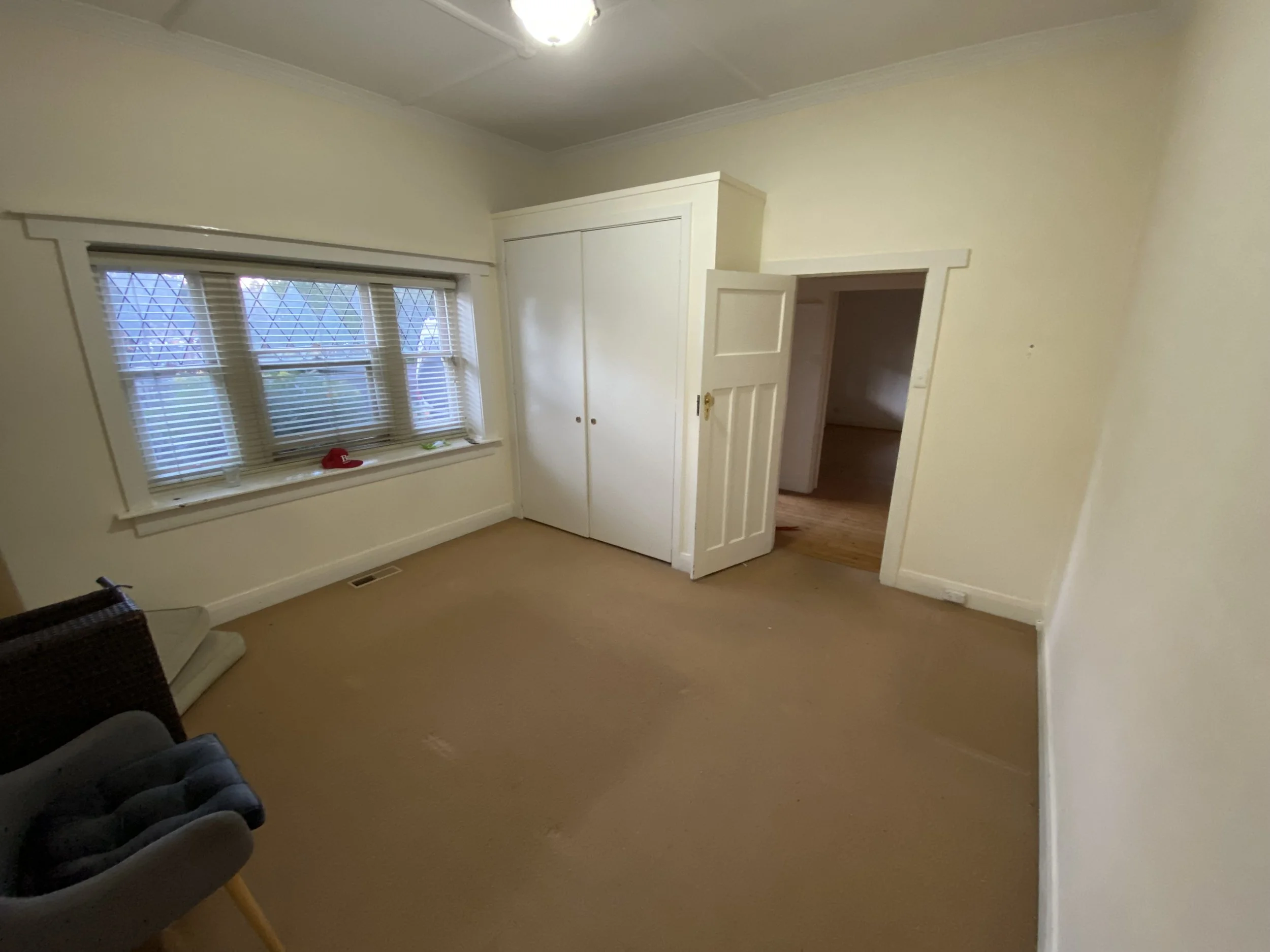 A bedroom with cream-colored walls, a window with blinds, a white closet, and an open door leading to another room. There is a black woven chair and a red baseball cap on the window sill.