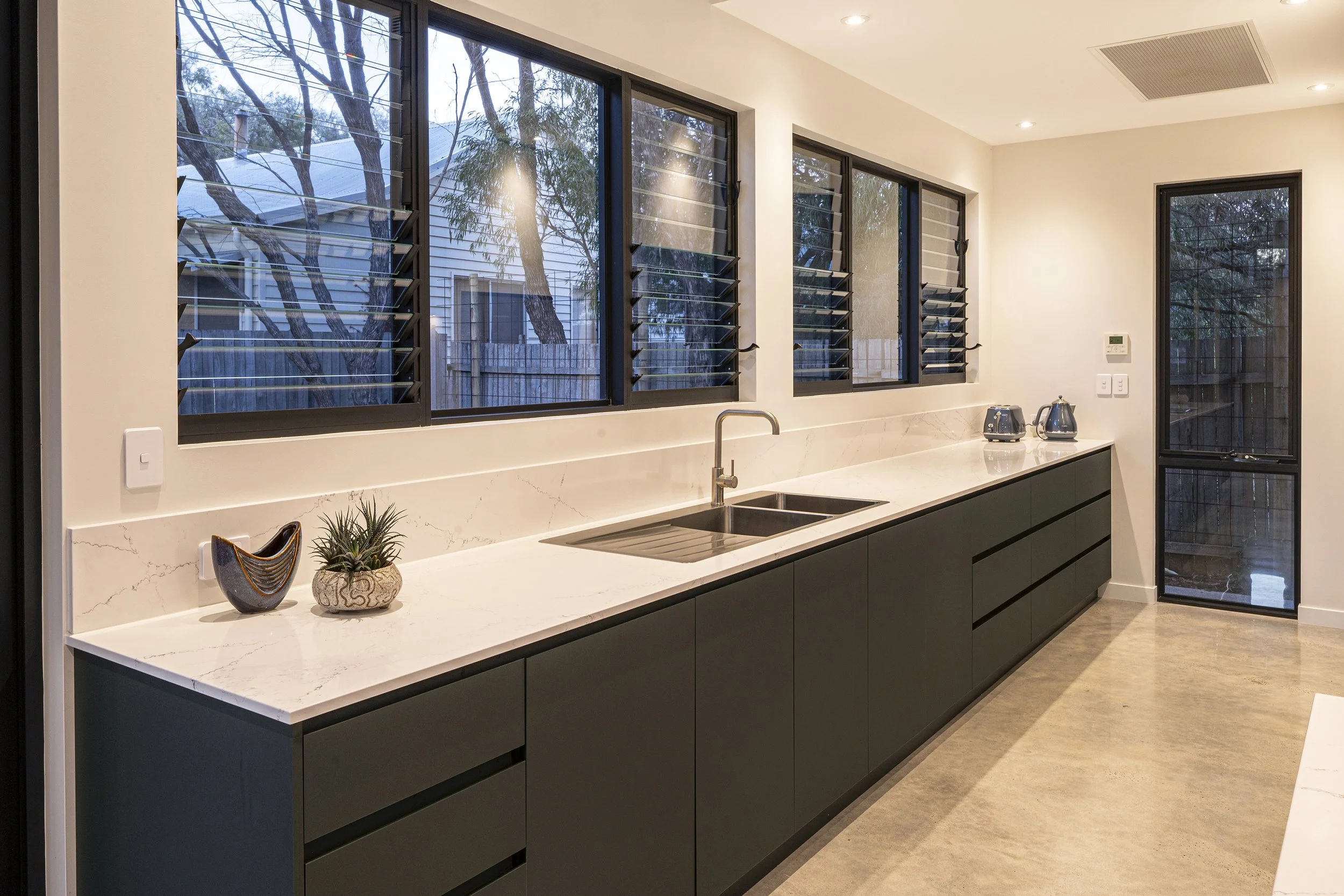 Modern kitchen with white marble countertop, black cabinets, large windows with adjustable louvered blinds, a sink, and small appliances including a toaster and kettle.