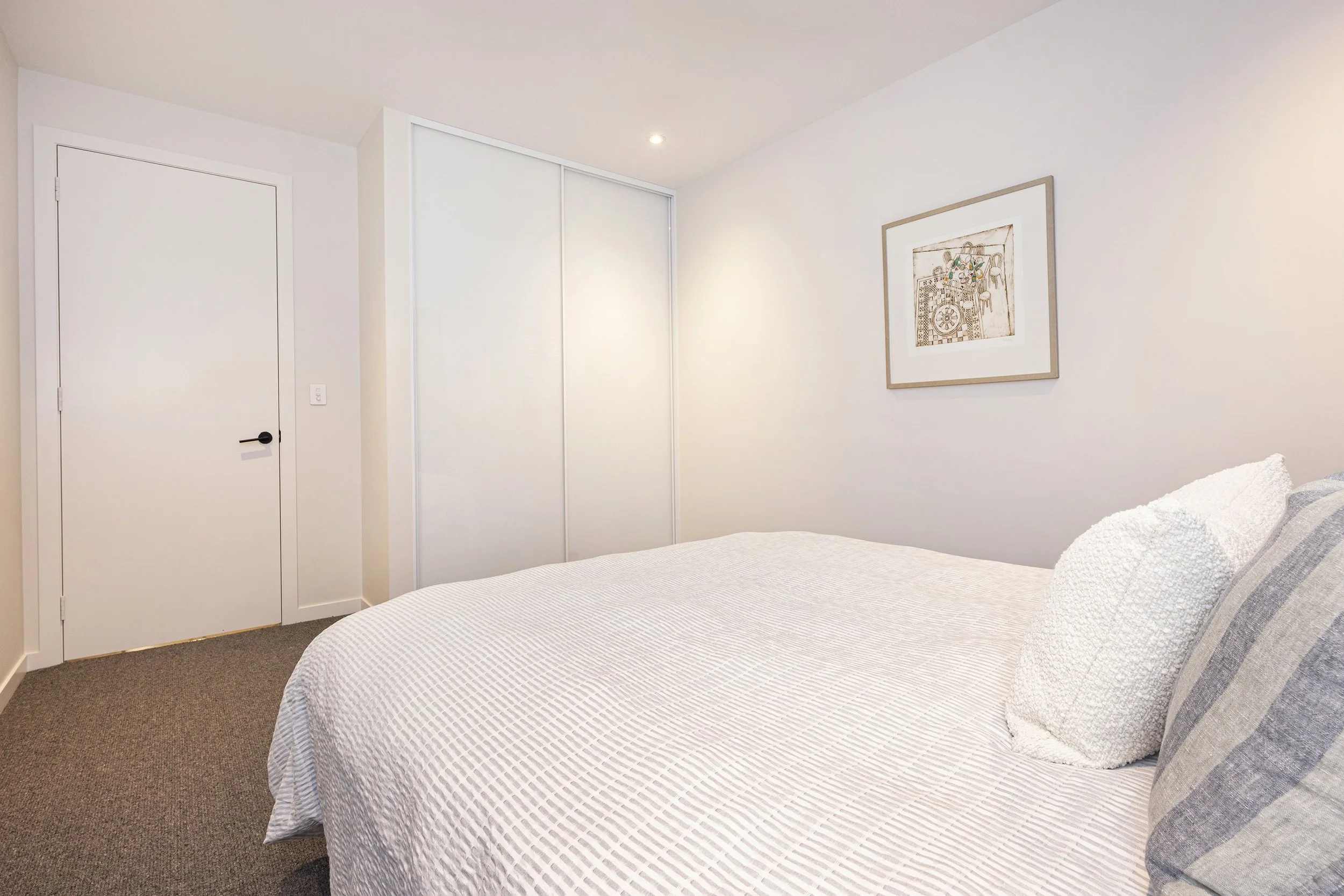 A bedroom with a bed covered in a striped white and gray bedspread, pillows, a framed artwork on the white wall, a closed door, and a sliding closet with white doors.
