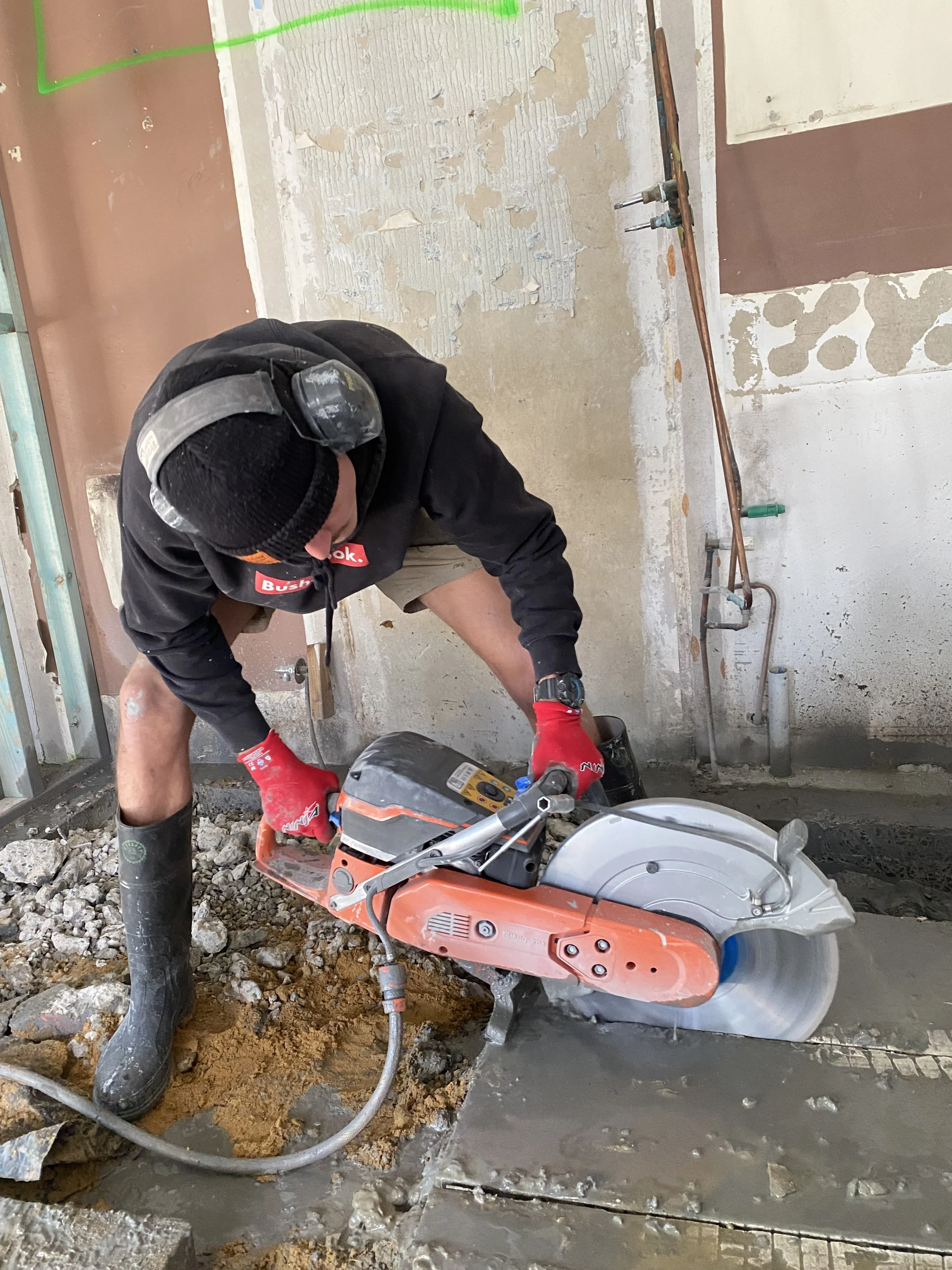 A construction worker wearing black clothing, red gloves, and black boots operating a concrete cutting saw at a construction site. The worker is kneeling on the floor surrounded by rocks and wet concrete, with partially peeled walls and construction 
