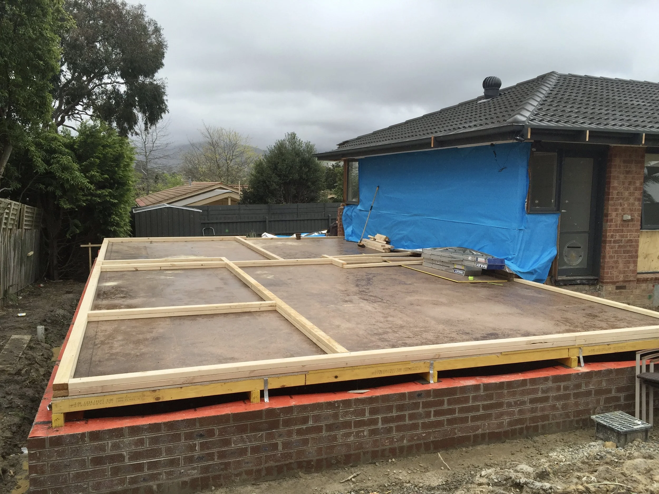 Construction site of a house, showing a brick foundation with some wooden framing and construction materials, with part of the house built featuring brick walls and a tiled roof, and a new elevated wooden platform in the foreground.
