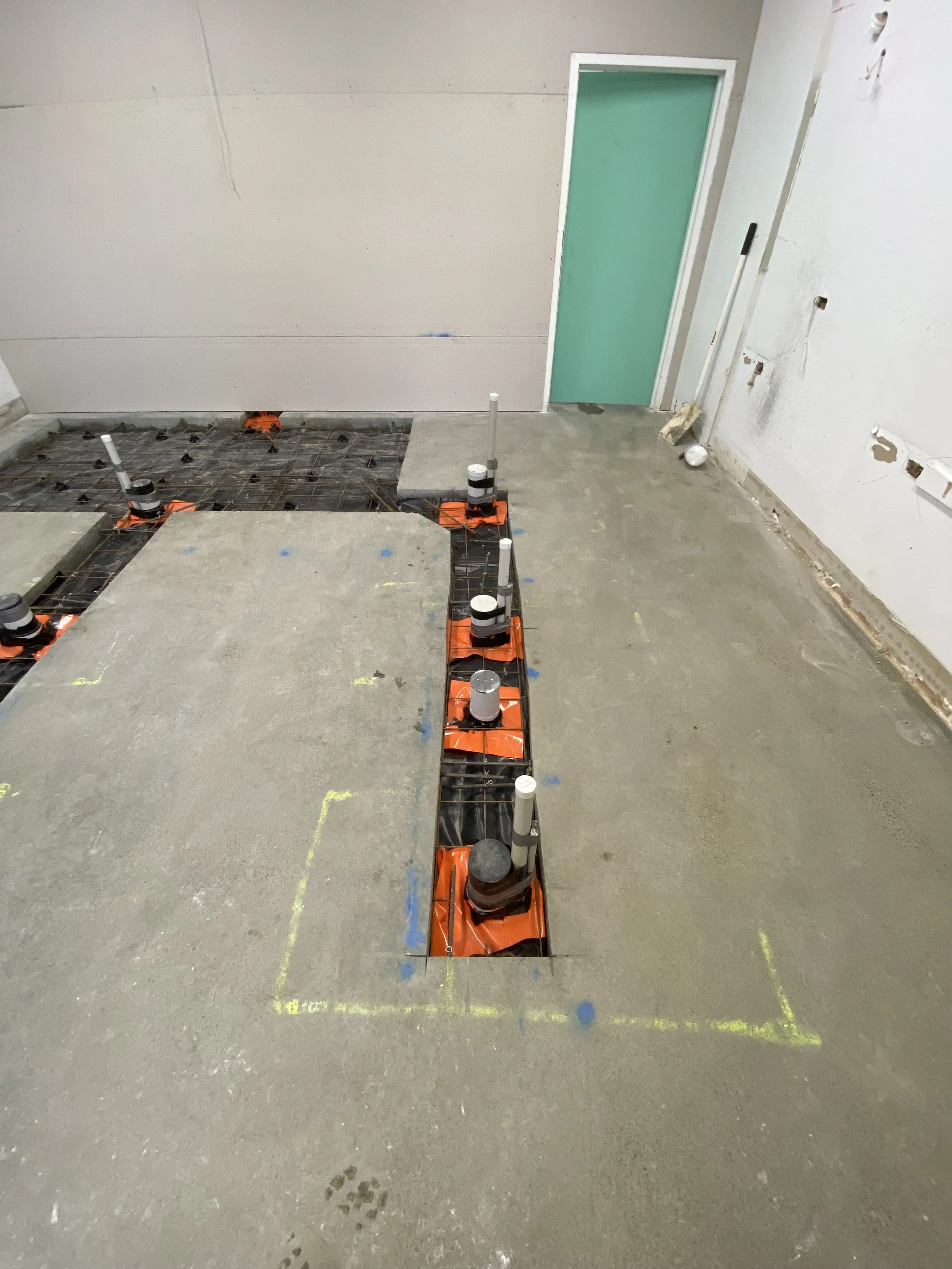 Construction site with a concrete floor, partially laid with a grid of rebar, and a green door on the wall. Several pipes are arranged on the floor, with some capped and others extending upward. There are utility boxes and pipes on the wall to the ri