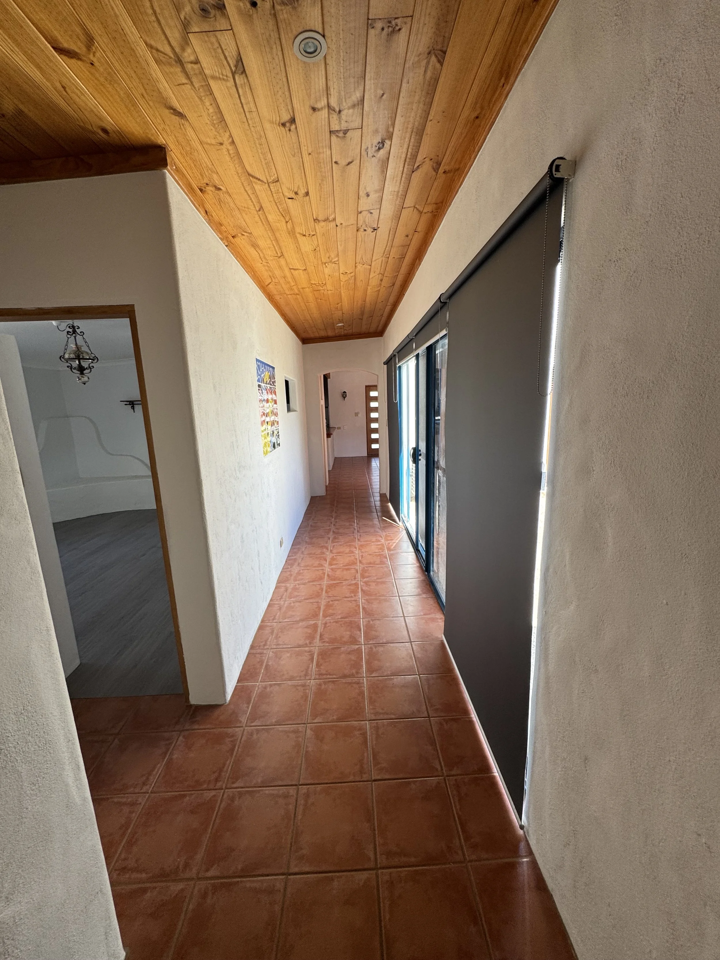 A hallway with a wooden ceiling, white textured walls, brown tiled floor, a sliding glass door with a blackout blind, and artwork on the left wall.