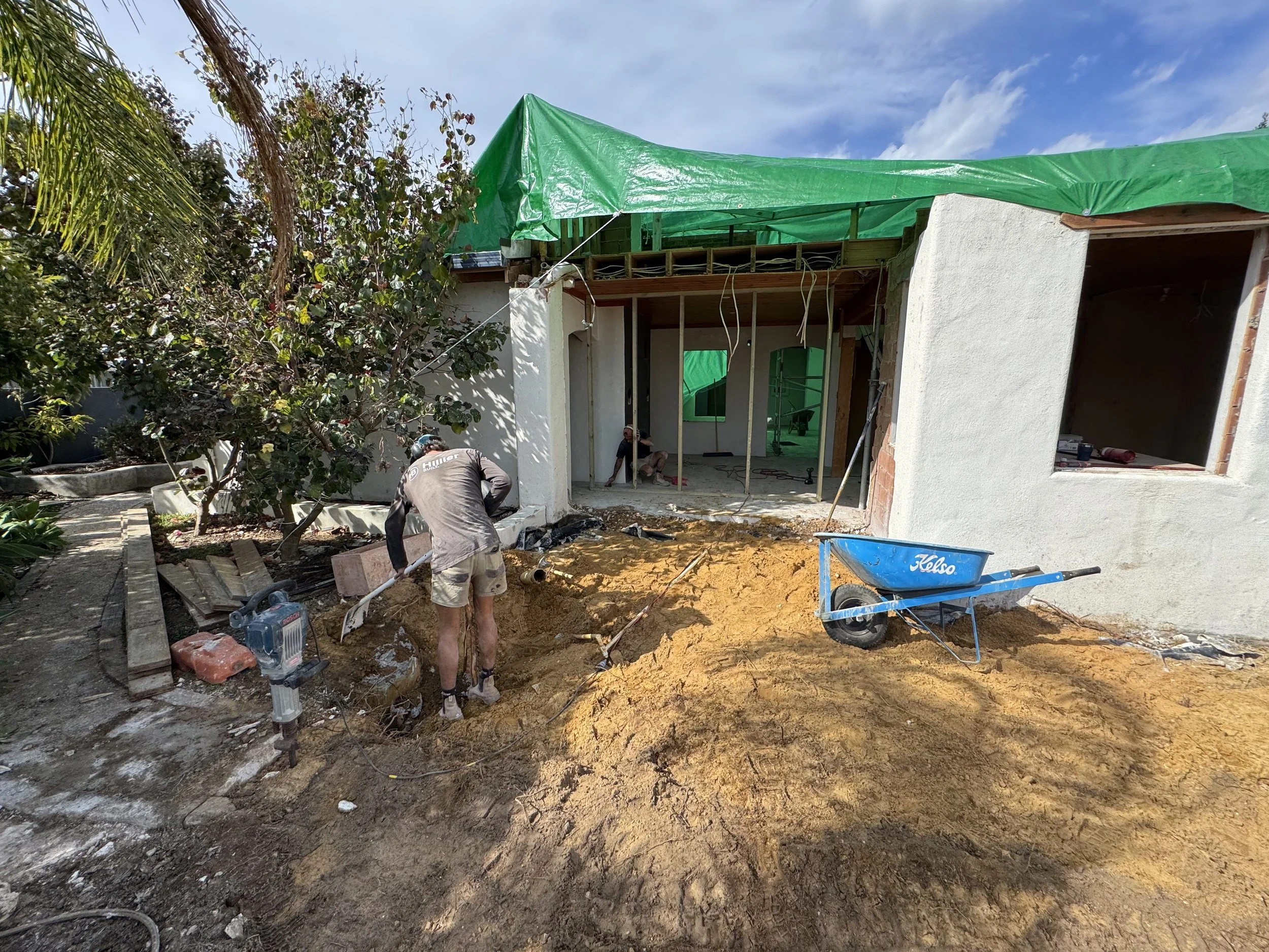 Construction workers working on a house renovation or extension, with a partially built wall and exposed wooden framing, surrounded by construction tools, materials, and dirt. Green scaffolding covers the upper part of the structure against a blue sk
