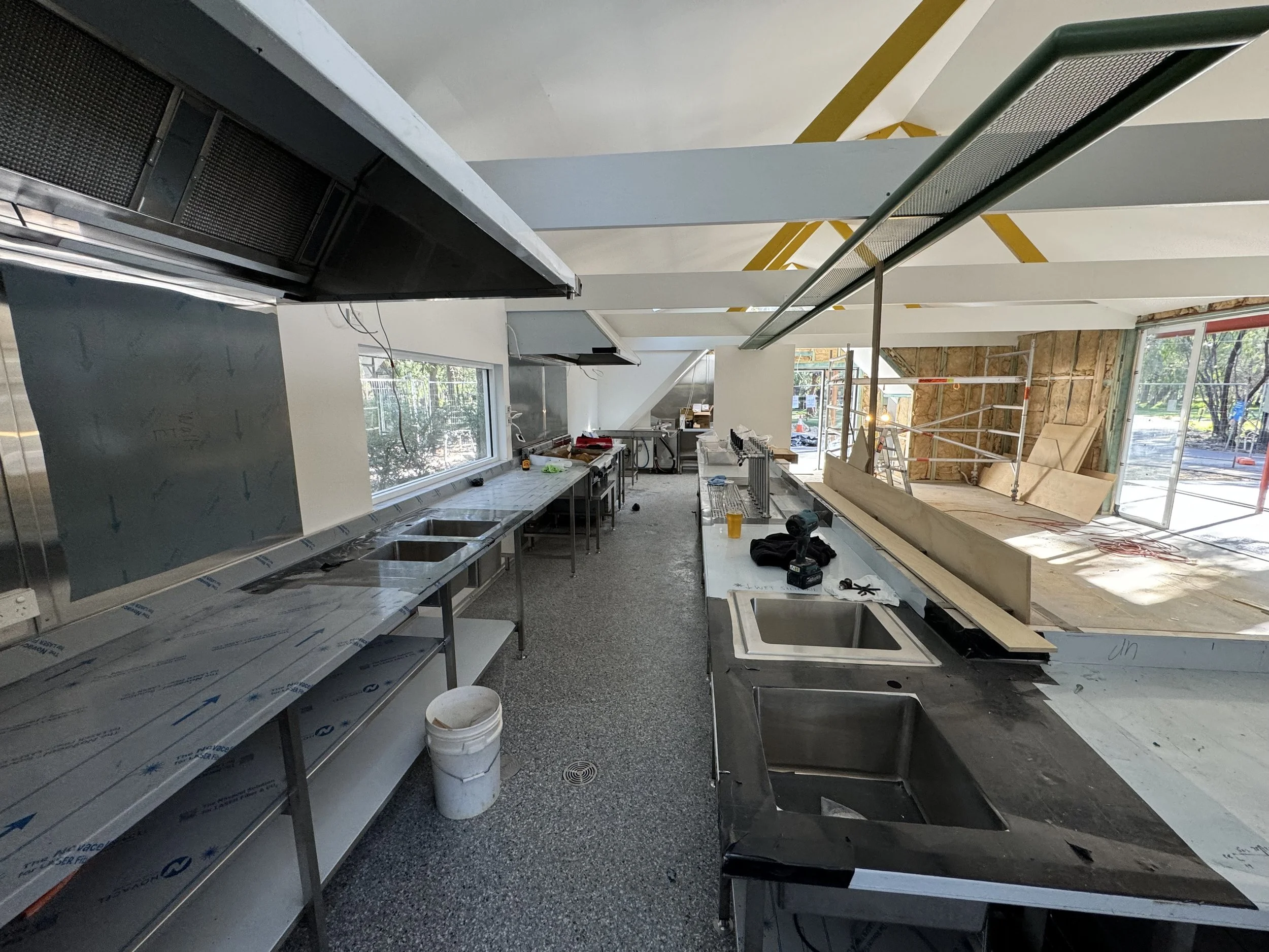 Interior of a commercial kitchen under construction, with stainless steel countertops, an industrial sink, and unfinished walls with exposed insulation and scaffolding.