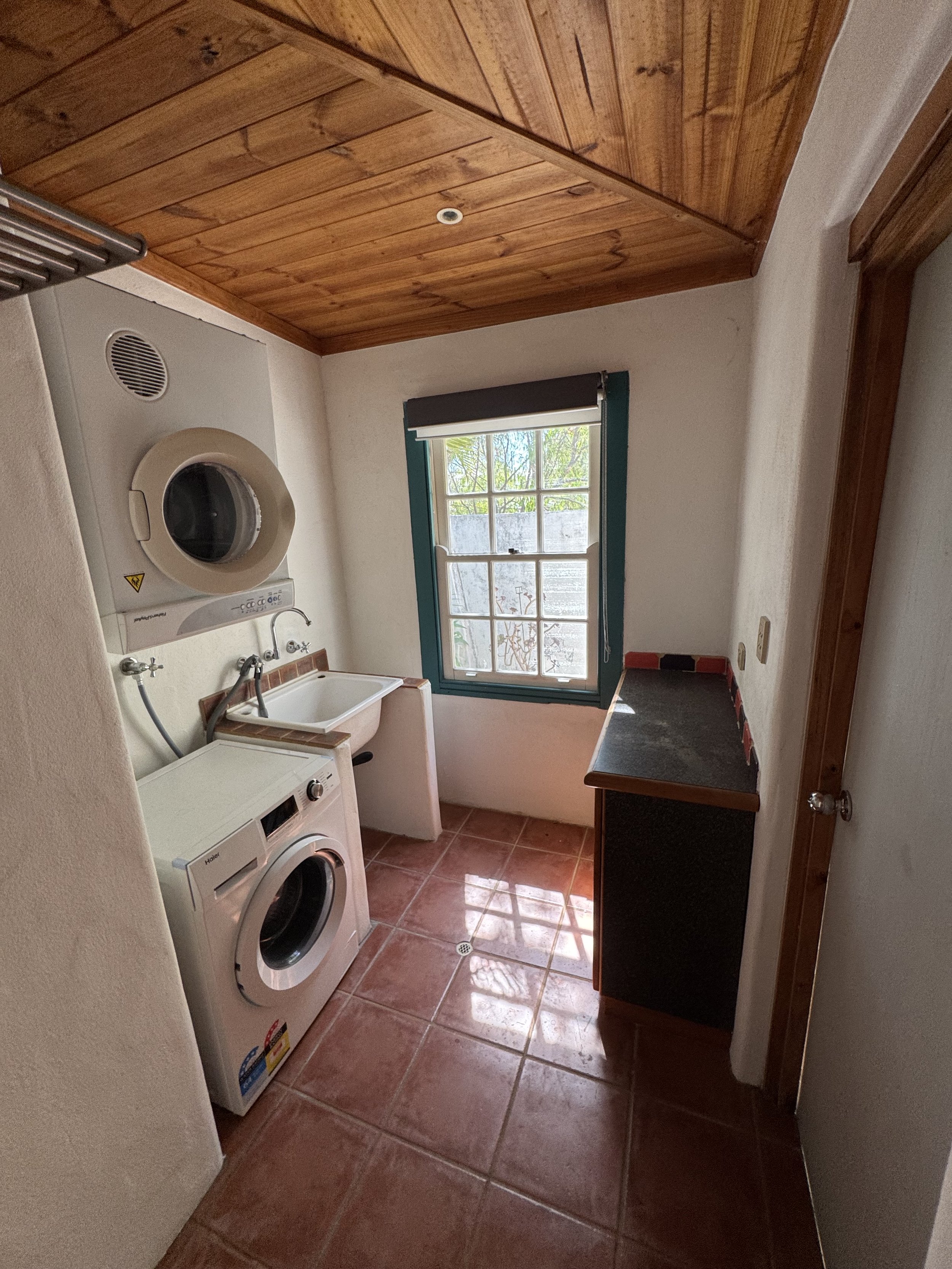 A small laundry room with a washer and dryer, a utility sink, a window with a view of greenery, and wooden ceiling paneling.