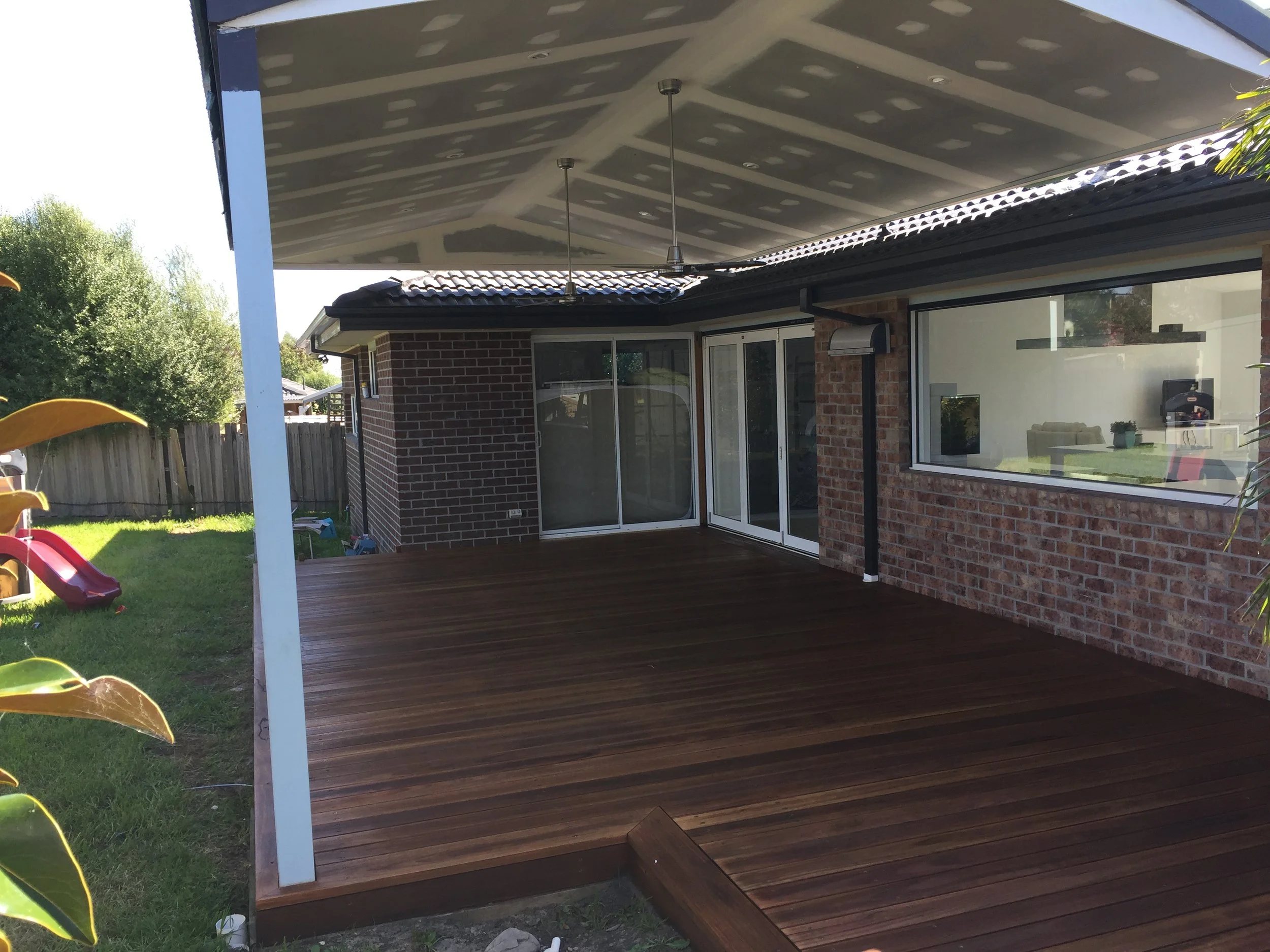 A covered outdoor patio with a wooden deck, attached to a brick house with large sliding glass doors. The roof of the patio is painted gray with multiple recessed lights, and there are two hanging pendant lights. The backyard has green grass, trees, 