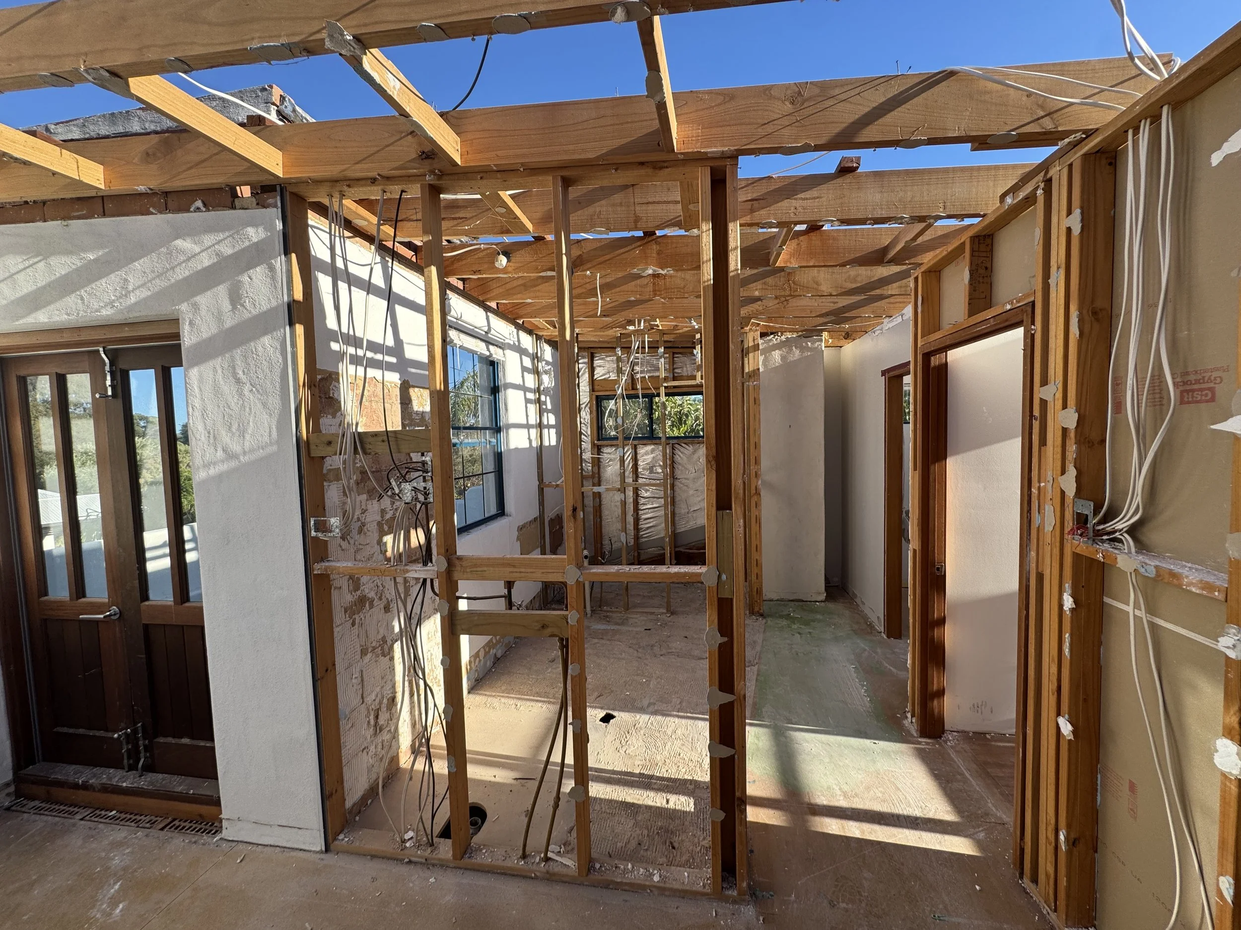 Interior of a house under renovation, with wooden framing and exposed ceiling beams, wiring, and unfinished walls.