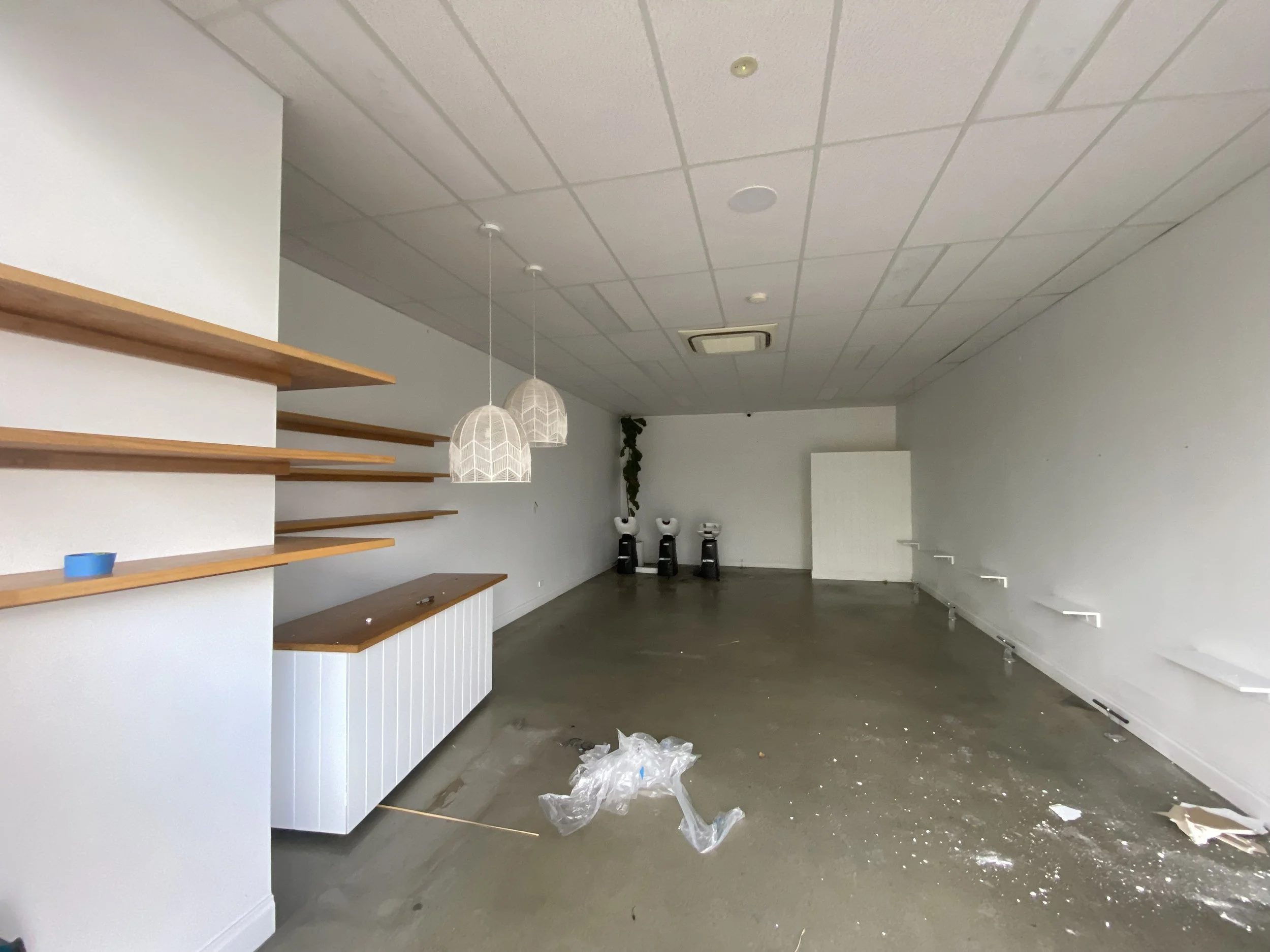 Empty interior with white walls, wooden shelves, hanging pendant lights, and chairs at the back. The floor is cluttered with plastic trash and debris.