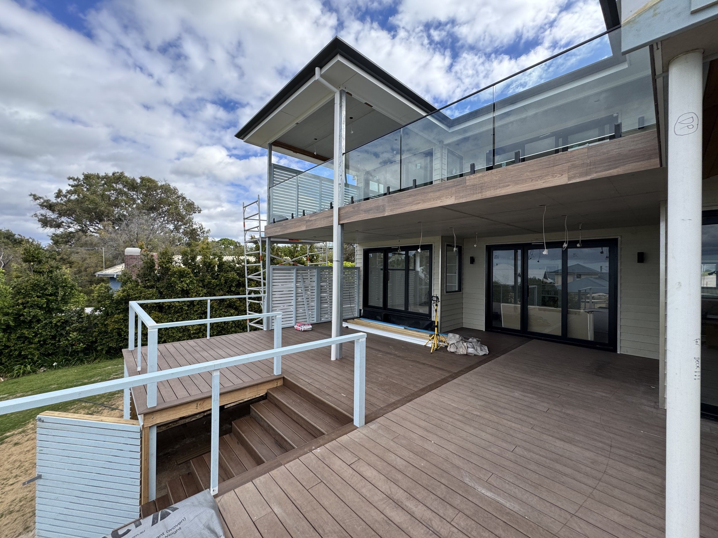 A house under construction with an outdoor wooden deck, glass railing, and scaffolding. The house has large sliding glass doors and a balcony above. The sky is partly cloudy, and there are trees in the background.