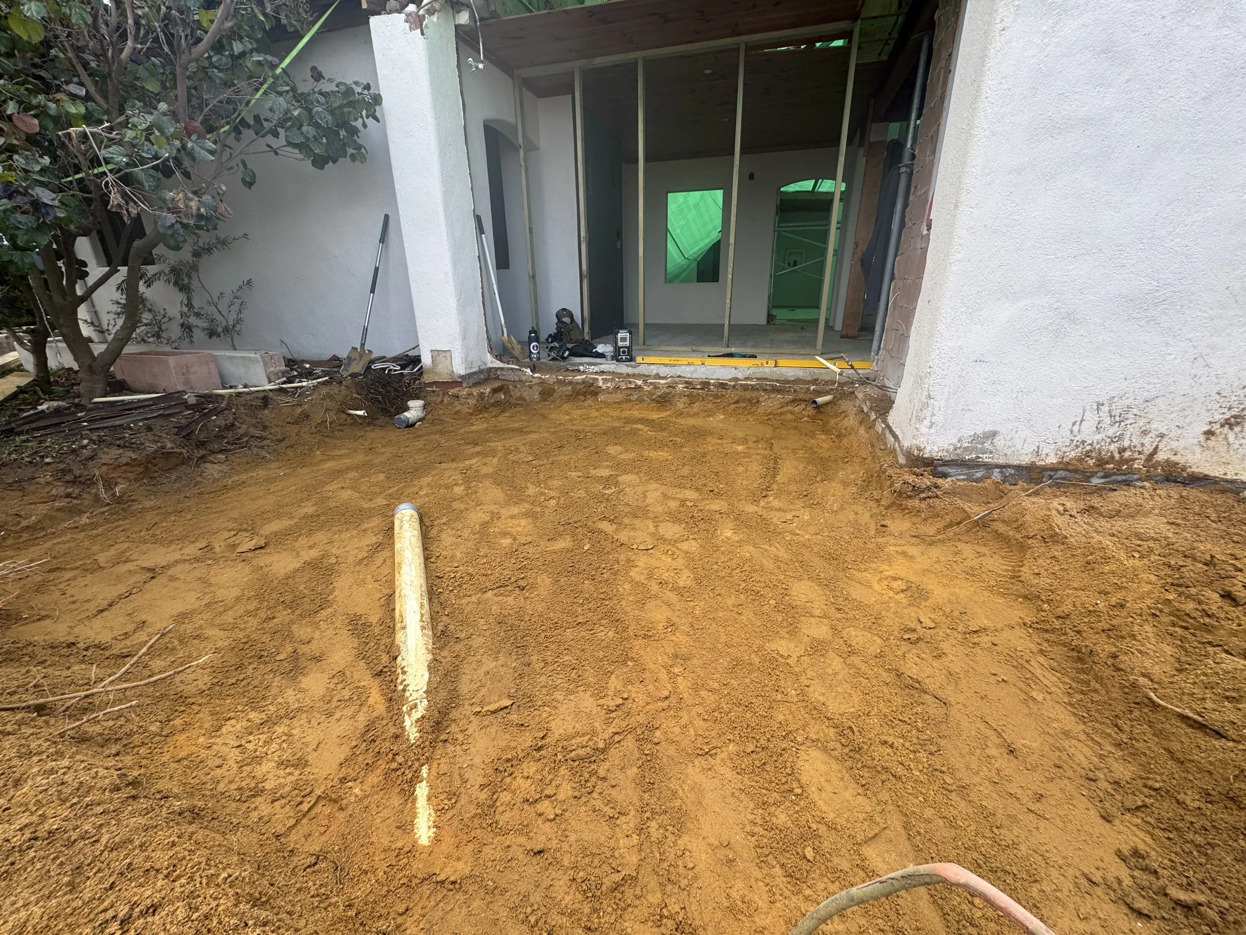 Construction site with a dug out area in front of a house, showing exposed soil, pipes, and construction tools.