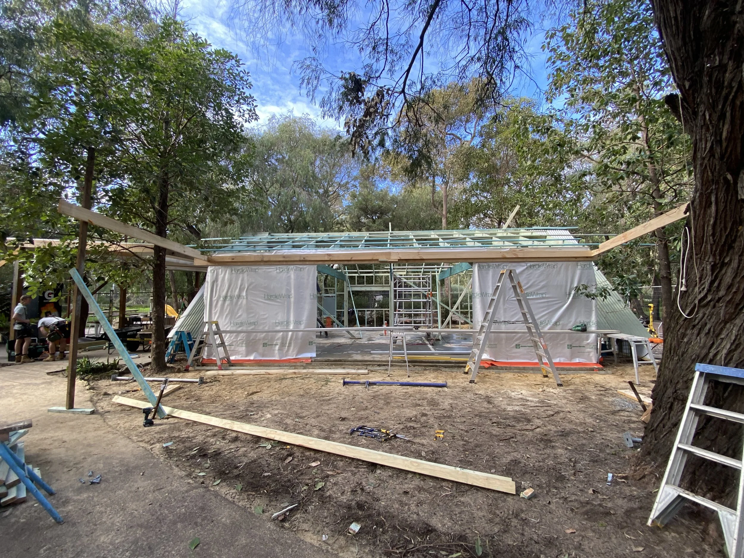 Construction site with a partially built roof structure, various ladders and tools, surrounded by trees and blue sky.