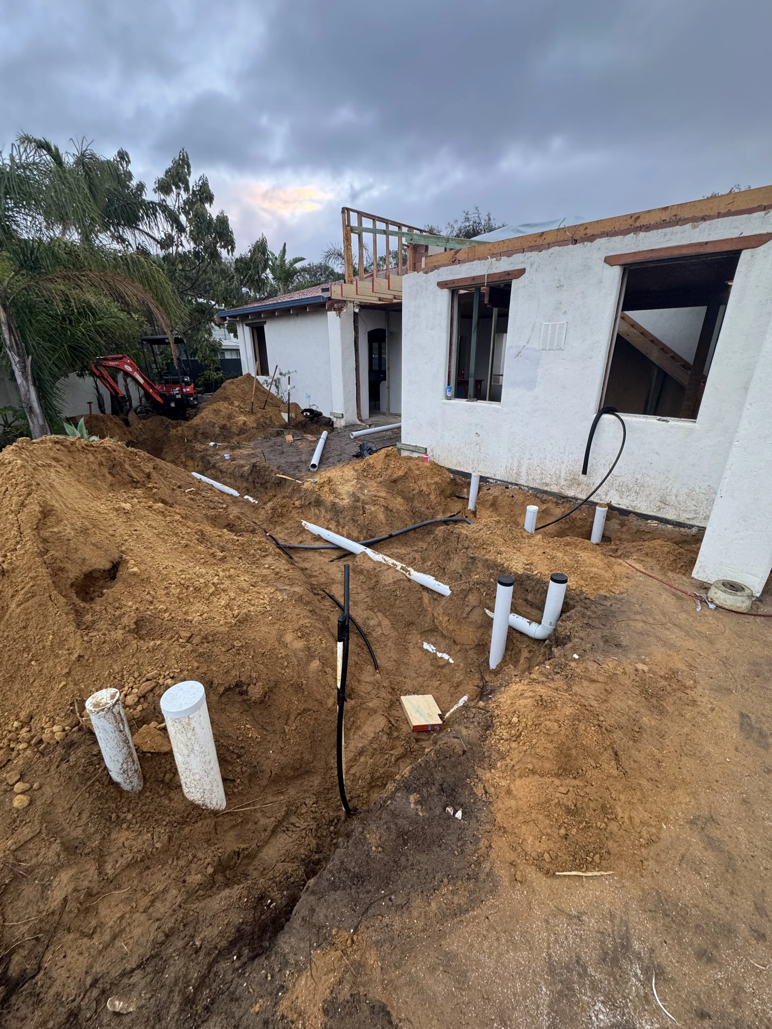 Construction site with excavated dirt, exposed pipes, and unfinished building walls with open window and door frames.