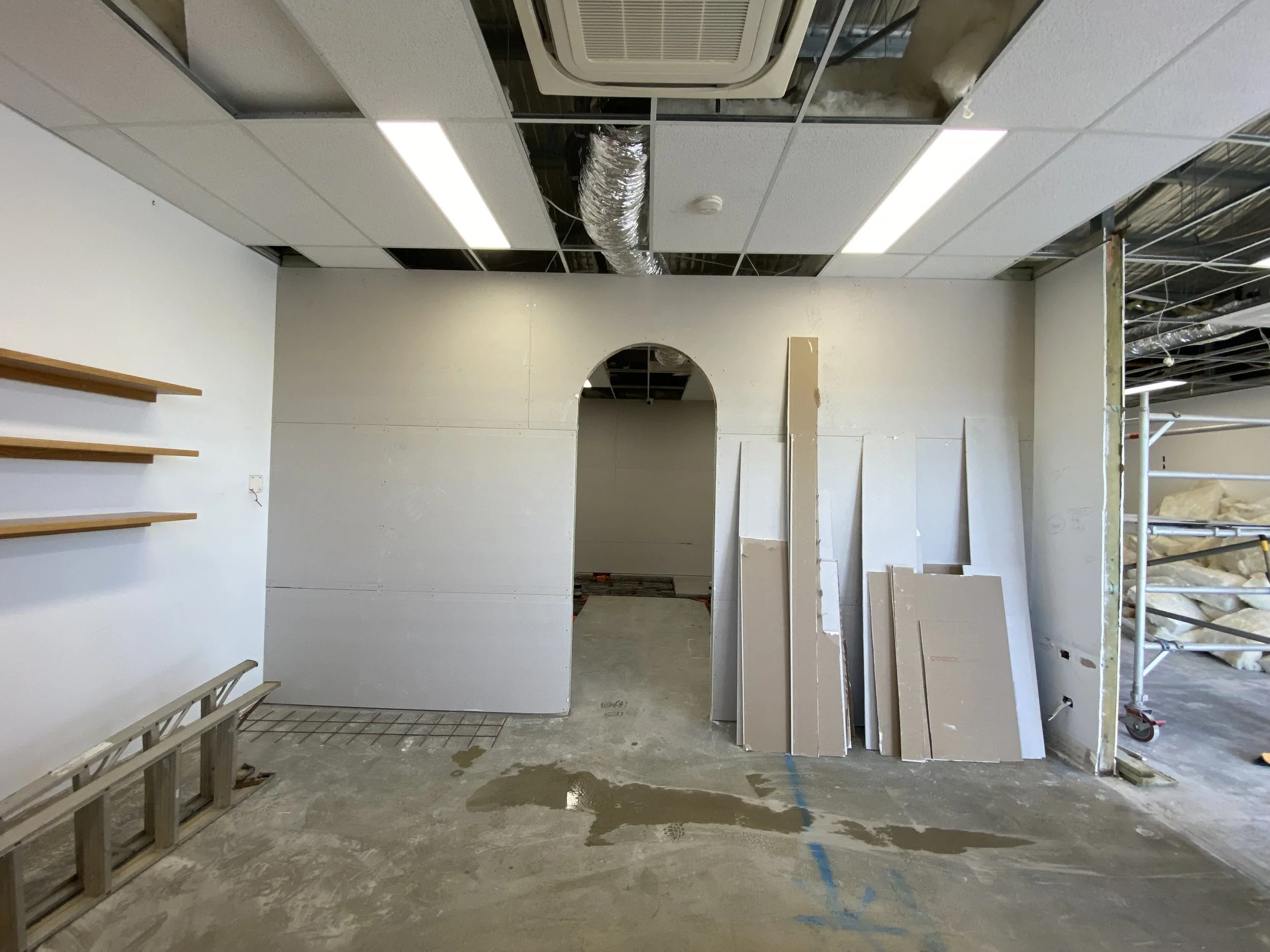 Interior of a building under construction with exposed ceiling ducts, an air conditioning unit, construction materials, and unfinished walls.