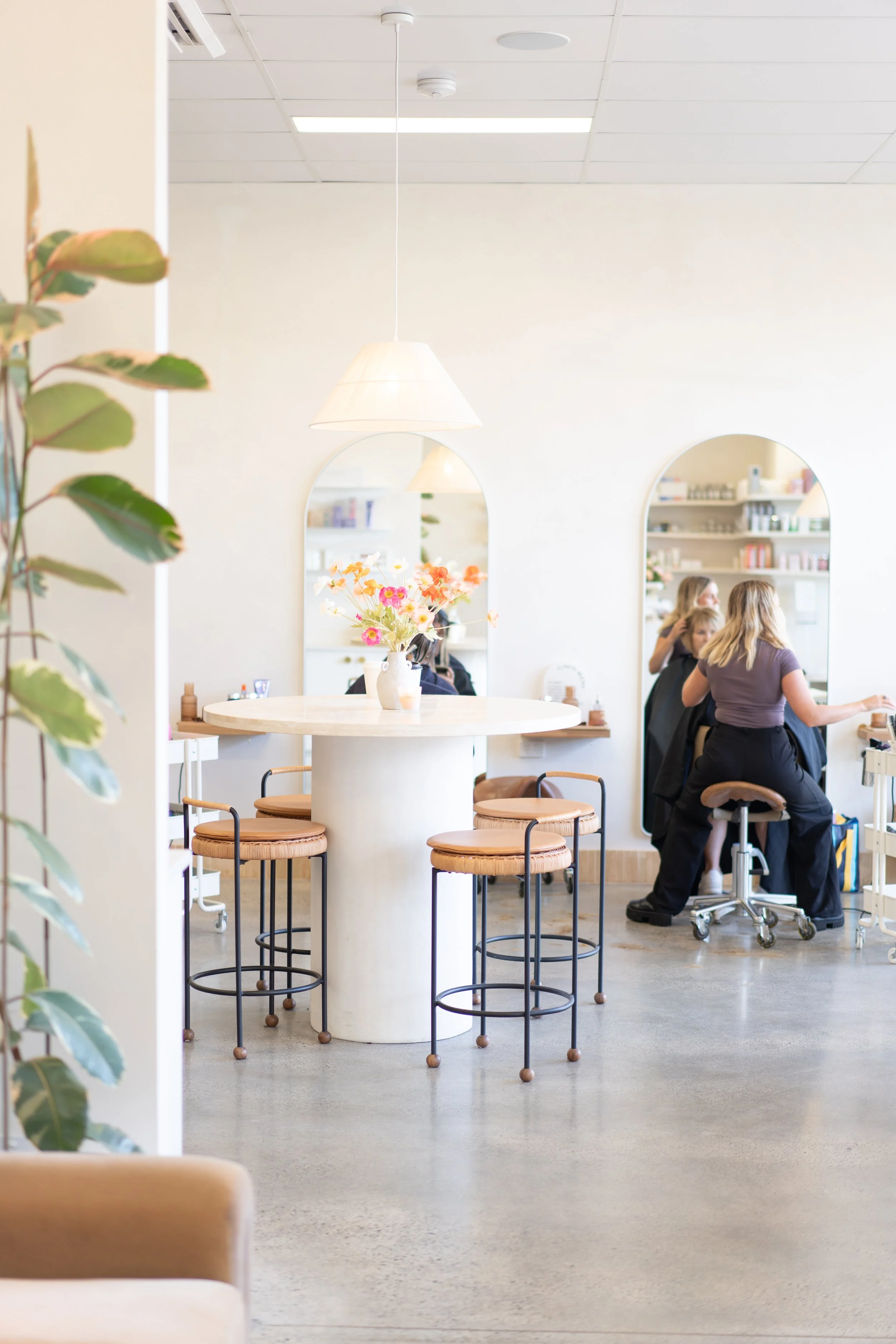 Interior of a modern, minimalist salon or beauty spa with a white round table, floral centerpiece, barstools, and two women receiving hair treatments.