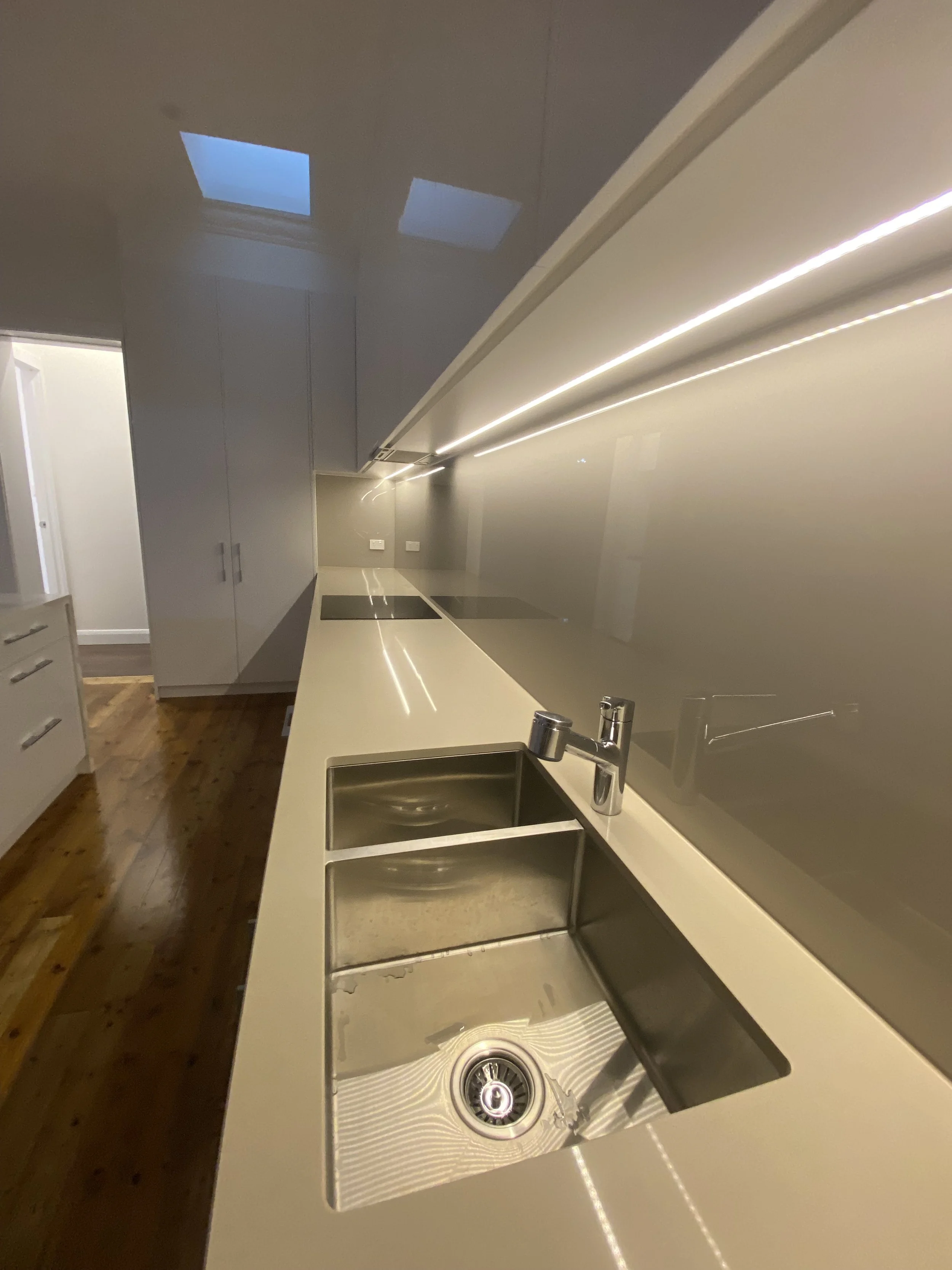 Modern kitchen with a beige countertop, stainless steel sink with double basins, black cutting board, and sleek cabinetry. Skylights on a celling provide natural light.