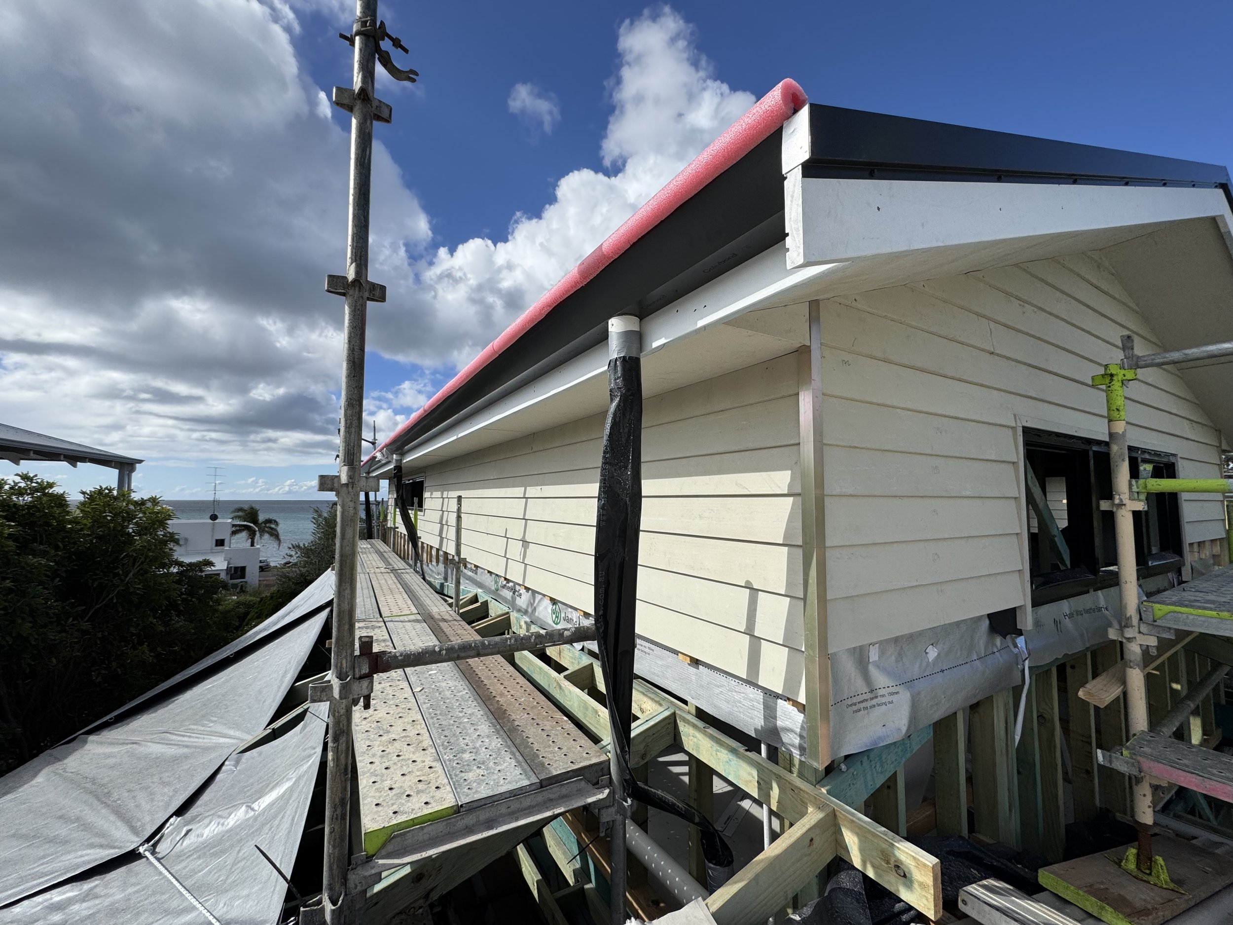 Construction scaffolding surrounding a house with light-colored siding, a partially installed roof edge with pink insulation foam, and a view of the sky with clouds and distant ocean.