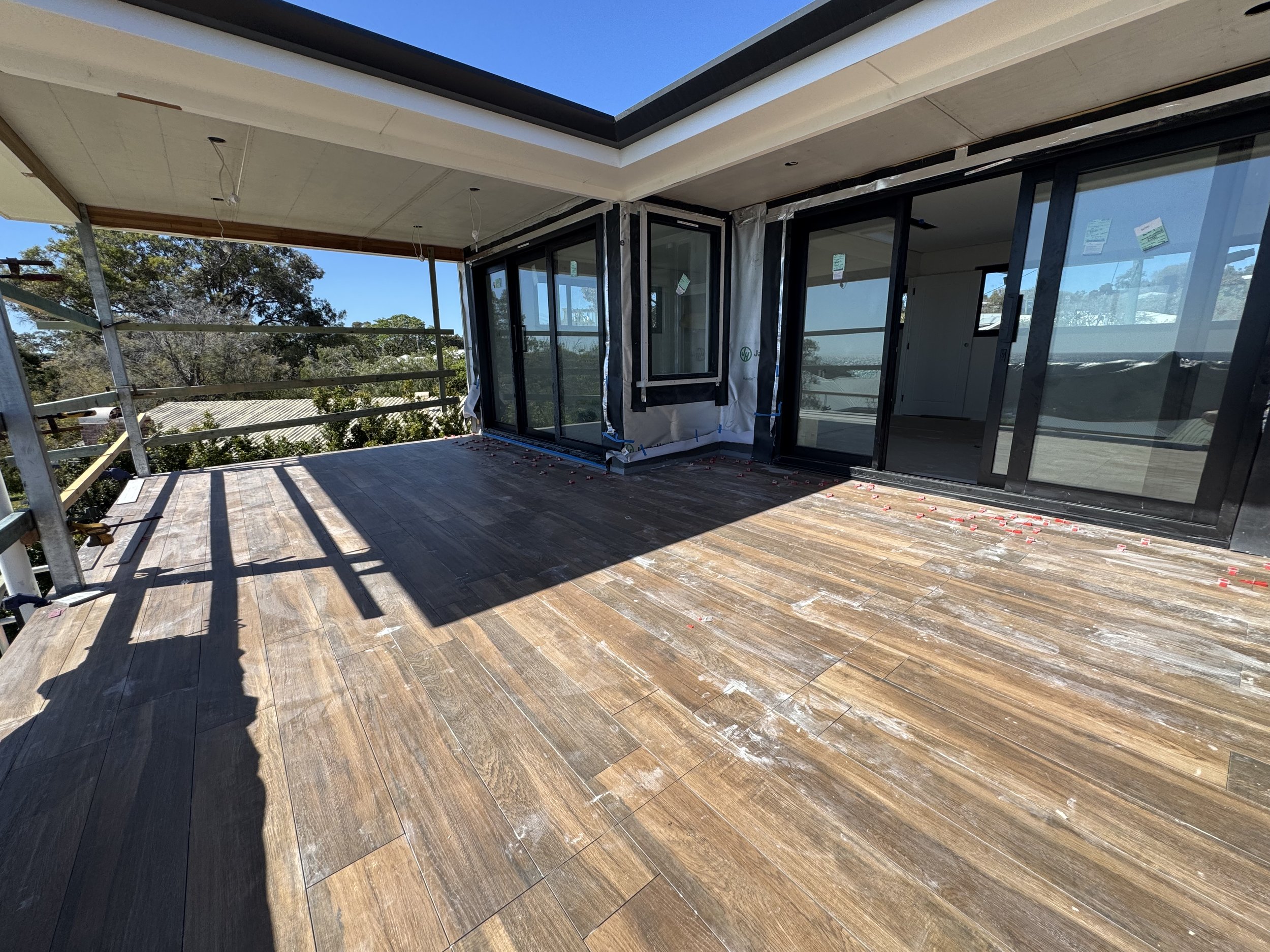 Under construction balcony with large sliding glass doors, wooden flooring, and partly finished ceiling, overlooking trees and a clear sky.
