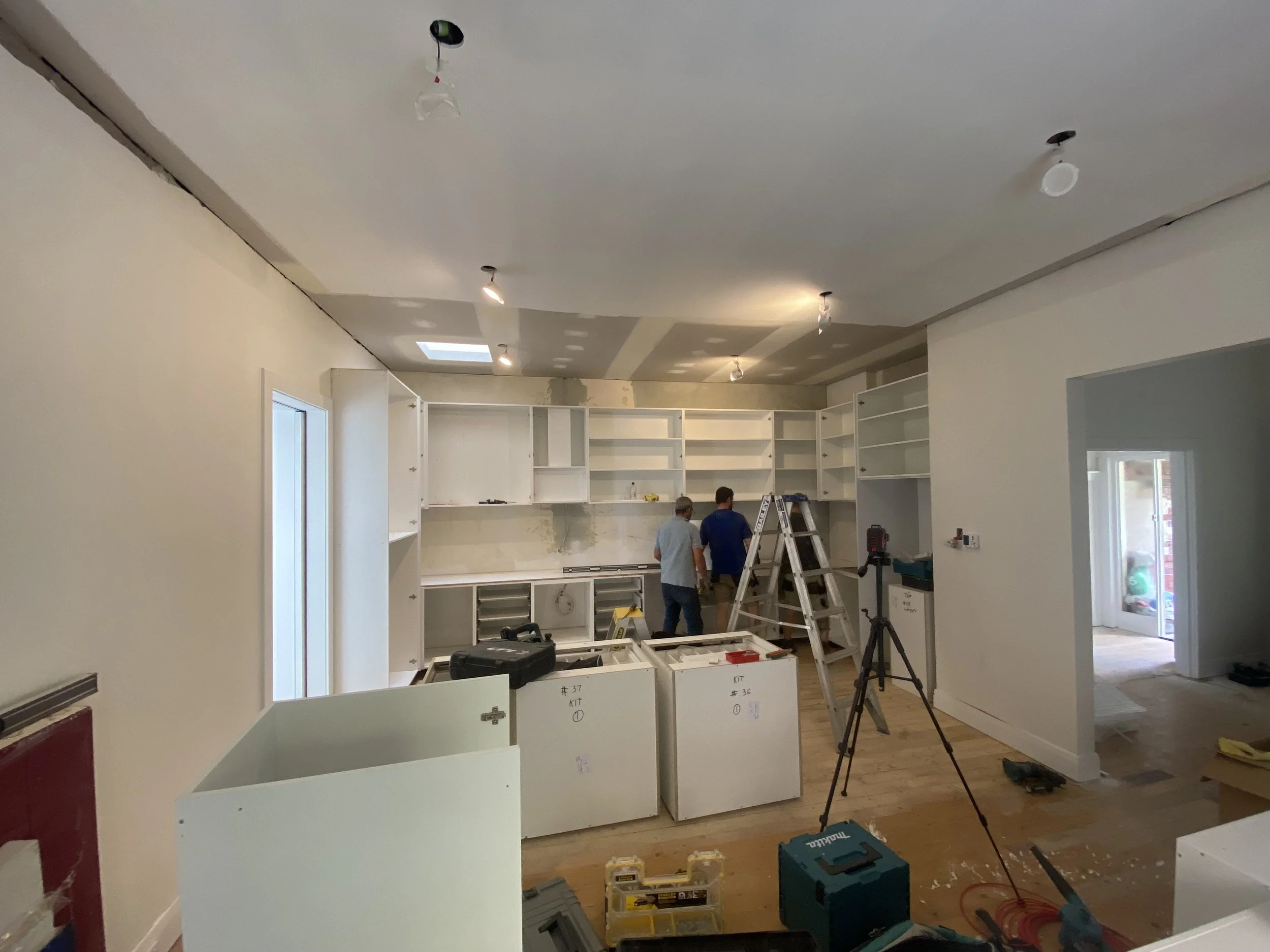 Construction workers installing white kitchen cabinets in a room with recessed lighting and a skylight, with tools and equipment scattered on the floor.