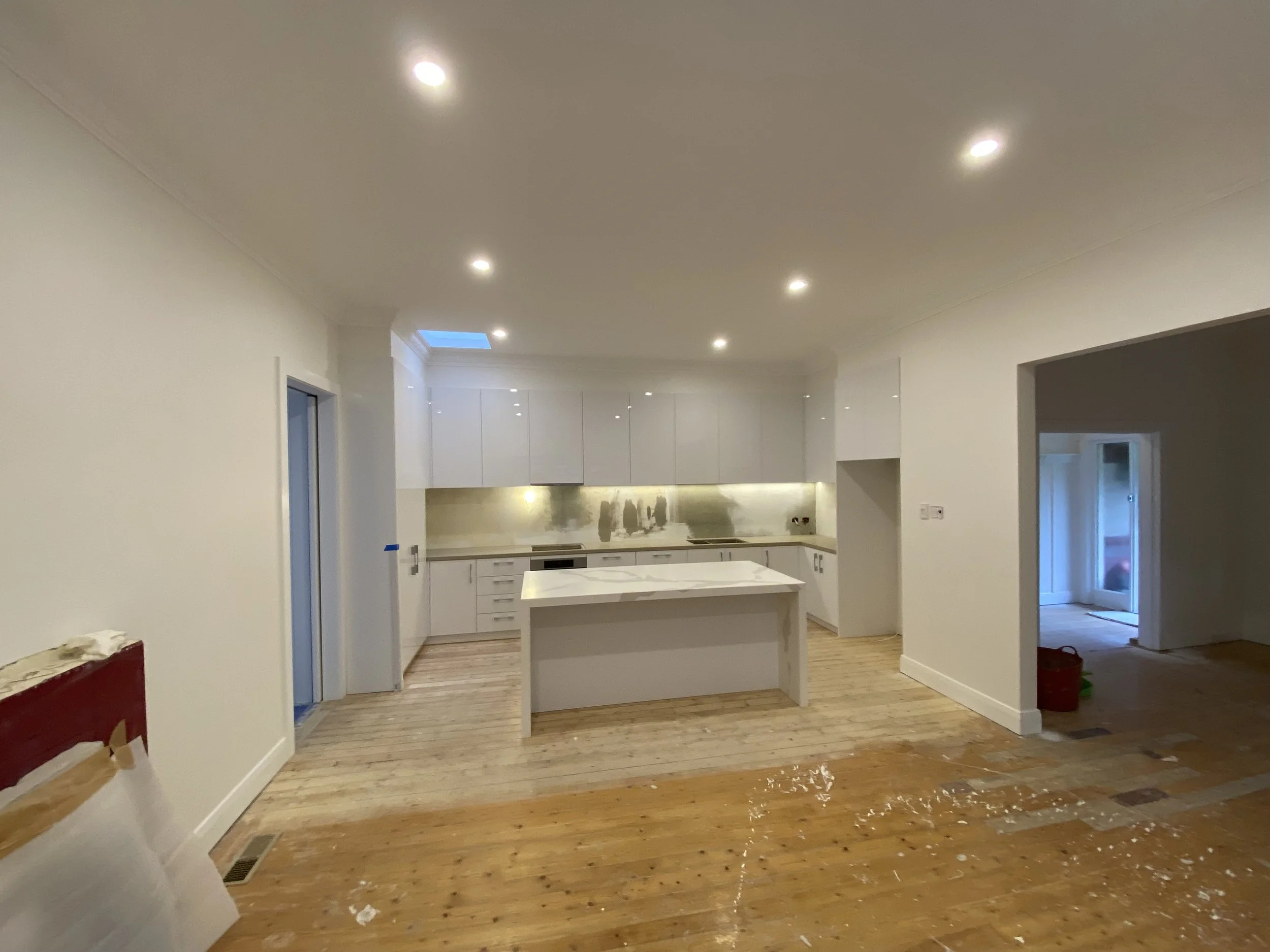 A spacious kitchen with white cabinets, a central island, and recessed ceiling lights. The floor appears to be wood, with some construction debris present, indicating ongoing renovation.