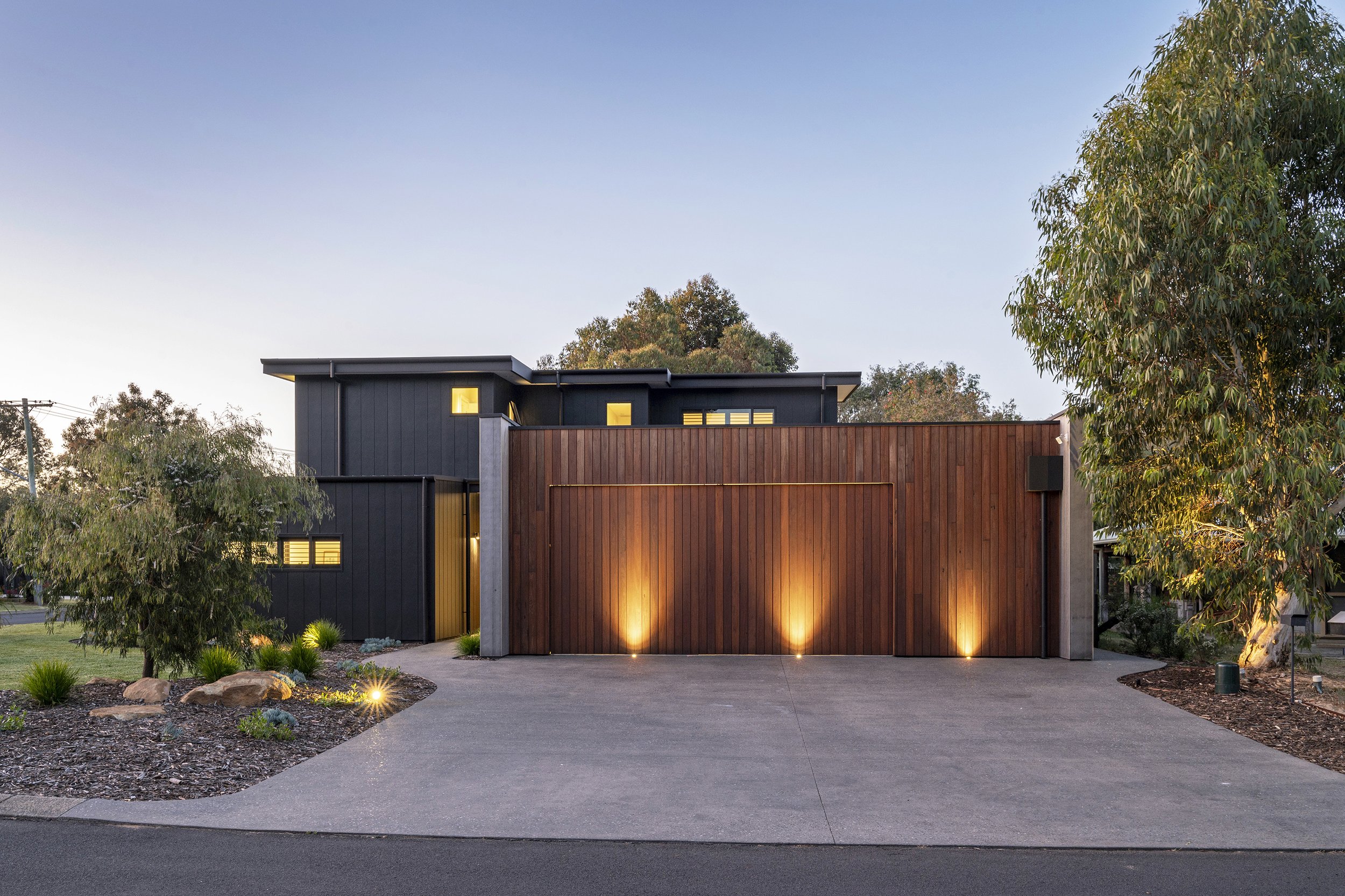 Modern two-story house with a wooden privacy fence, illuminated landscape lighting, surrounded by trees, under a clear evening sky.