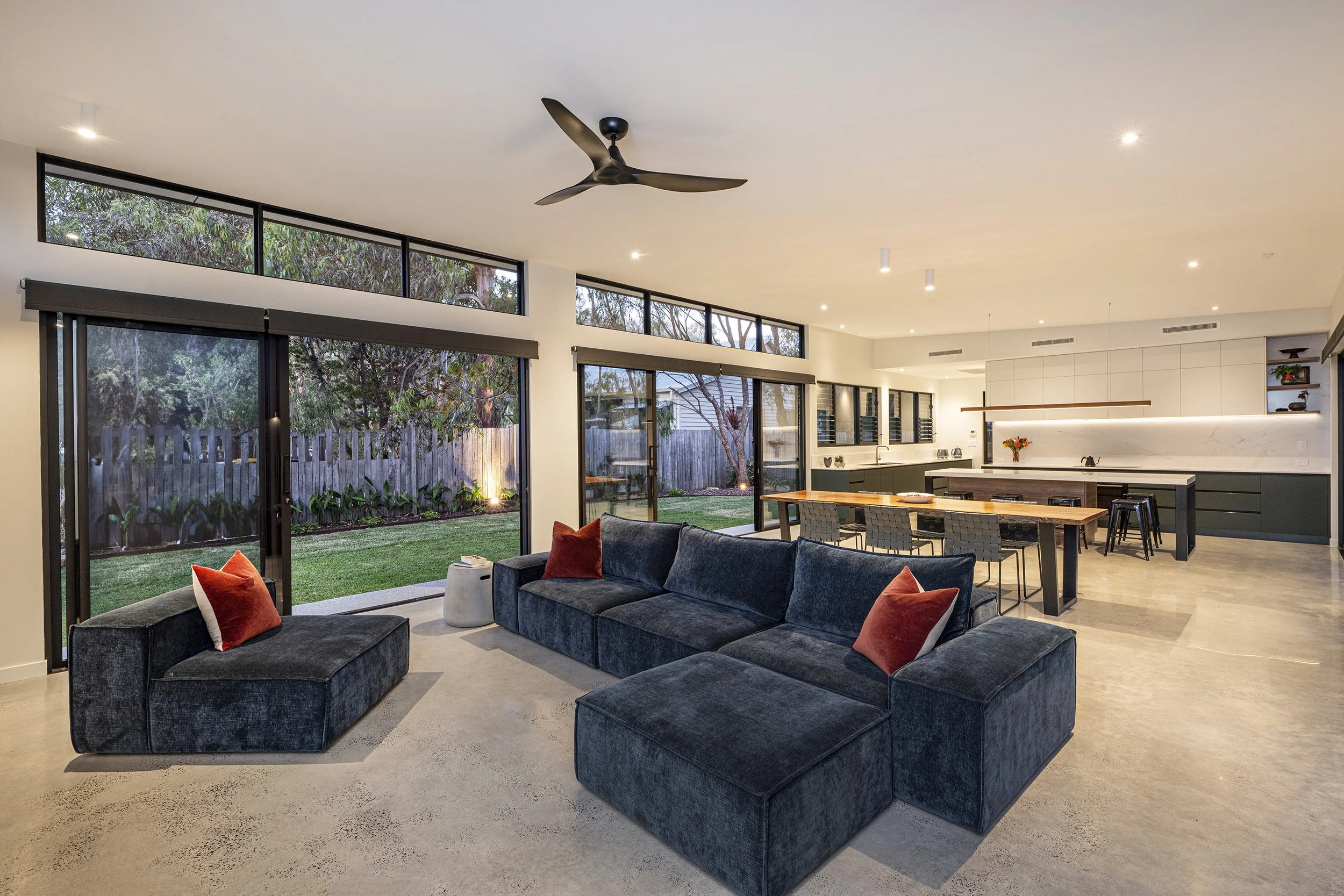Open-concept modern living room and kitchen with large windows and sliding doors opening to backyard with trees and a wooden fence, featuring a black ceiling fan, gray and blue sofas with orange pillows, a wooden dining table, and minimalist decor.