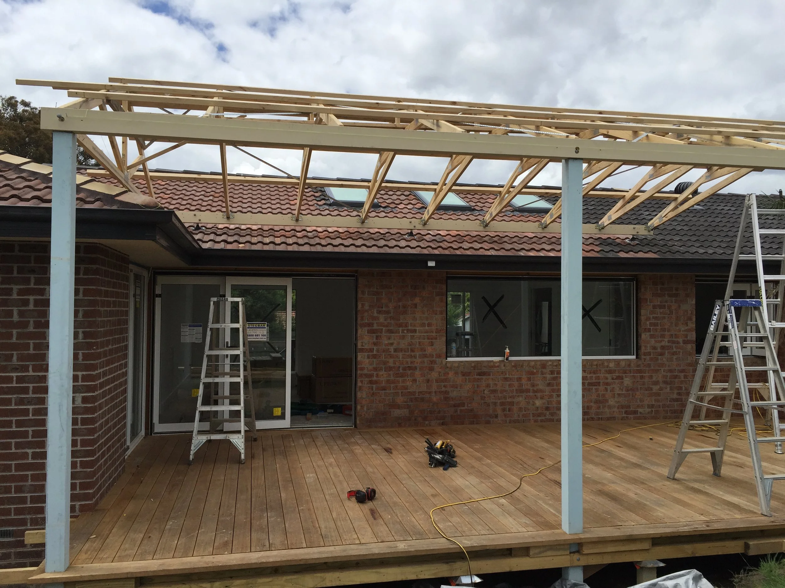 Construction of a wooden deck extension on the back of a brick house with a red tiled roof, ladders, tools, and building materials present.