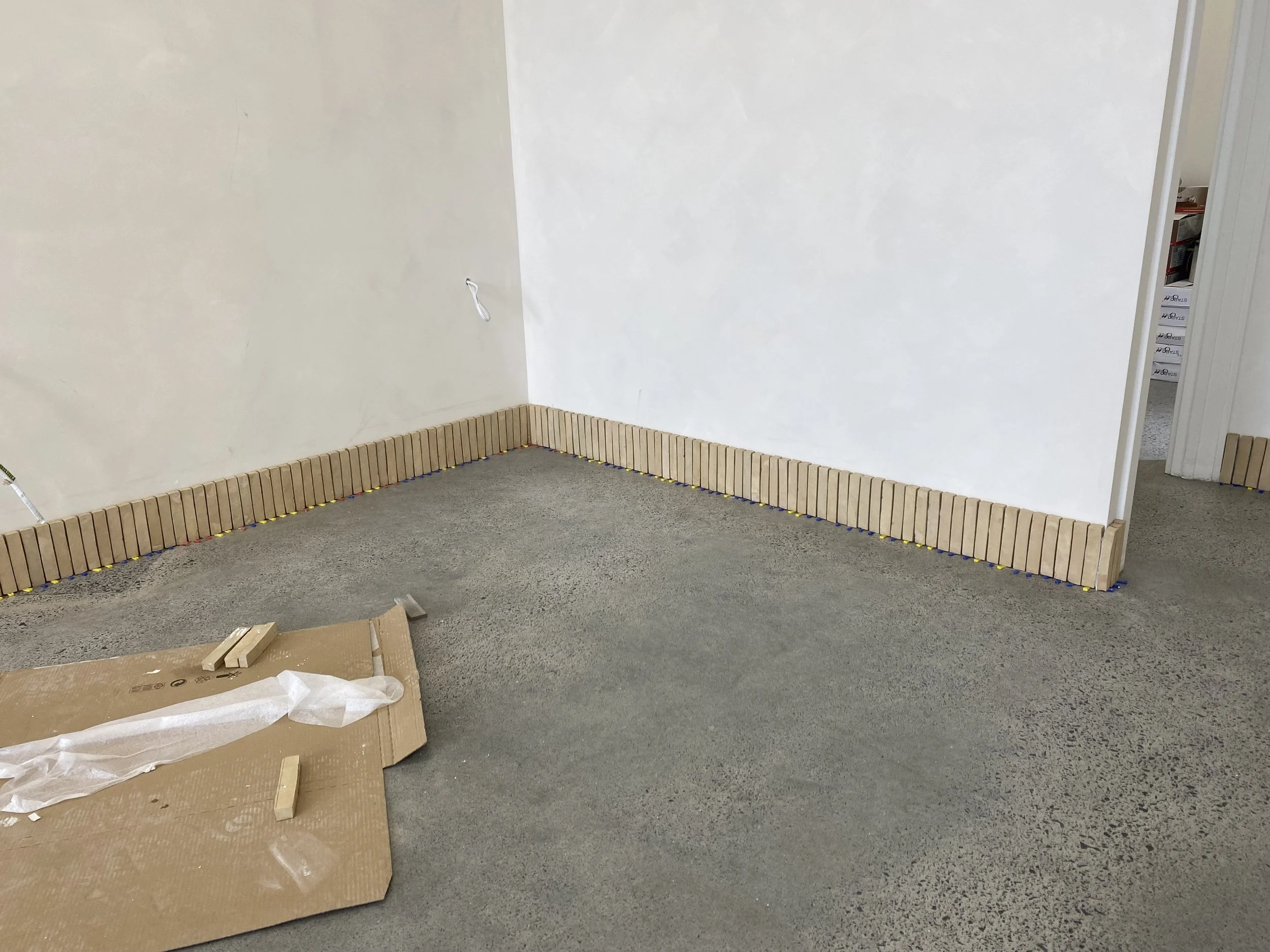 A corner of a room with white walls and a gray carpeted floor, where a border of small wooden blocks is being installed along the base of the walls.