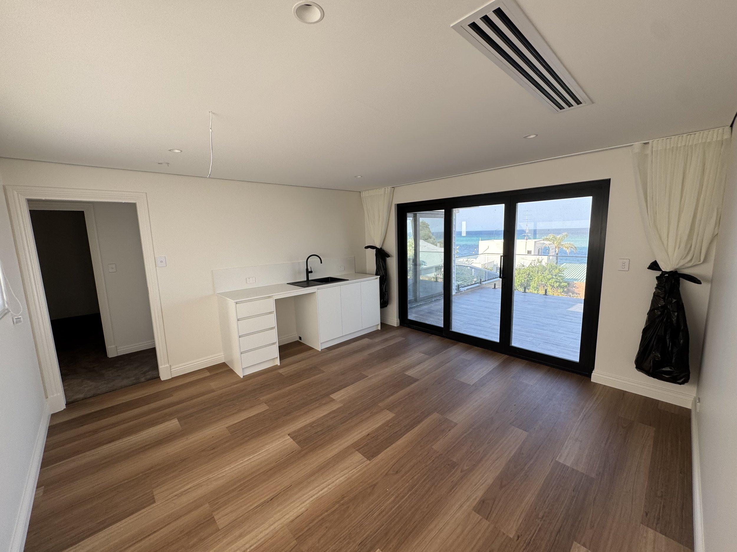 Empty living room with large sliding glass door showing a view of the ocean, with curtains tied back and hardwood floors.