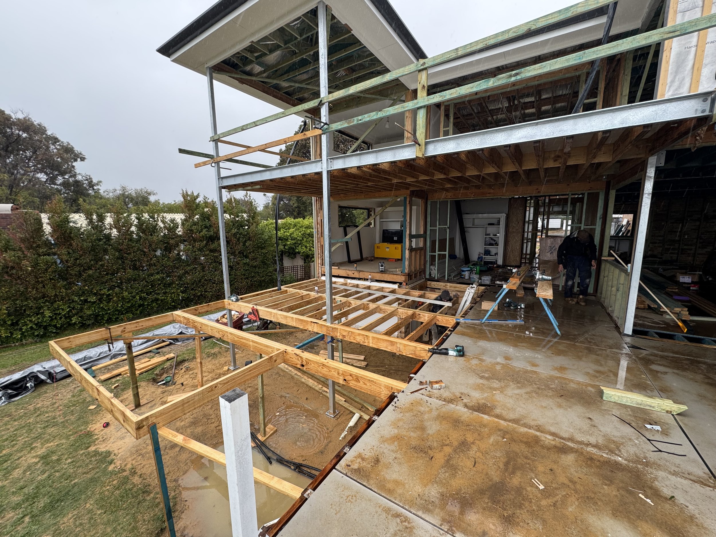 Construction site with a partially built multi-story house, showing wooden framing, metal supports, and construction tools. Overcast weather with rain, and trees in the background.