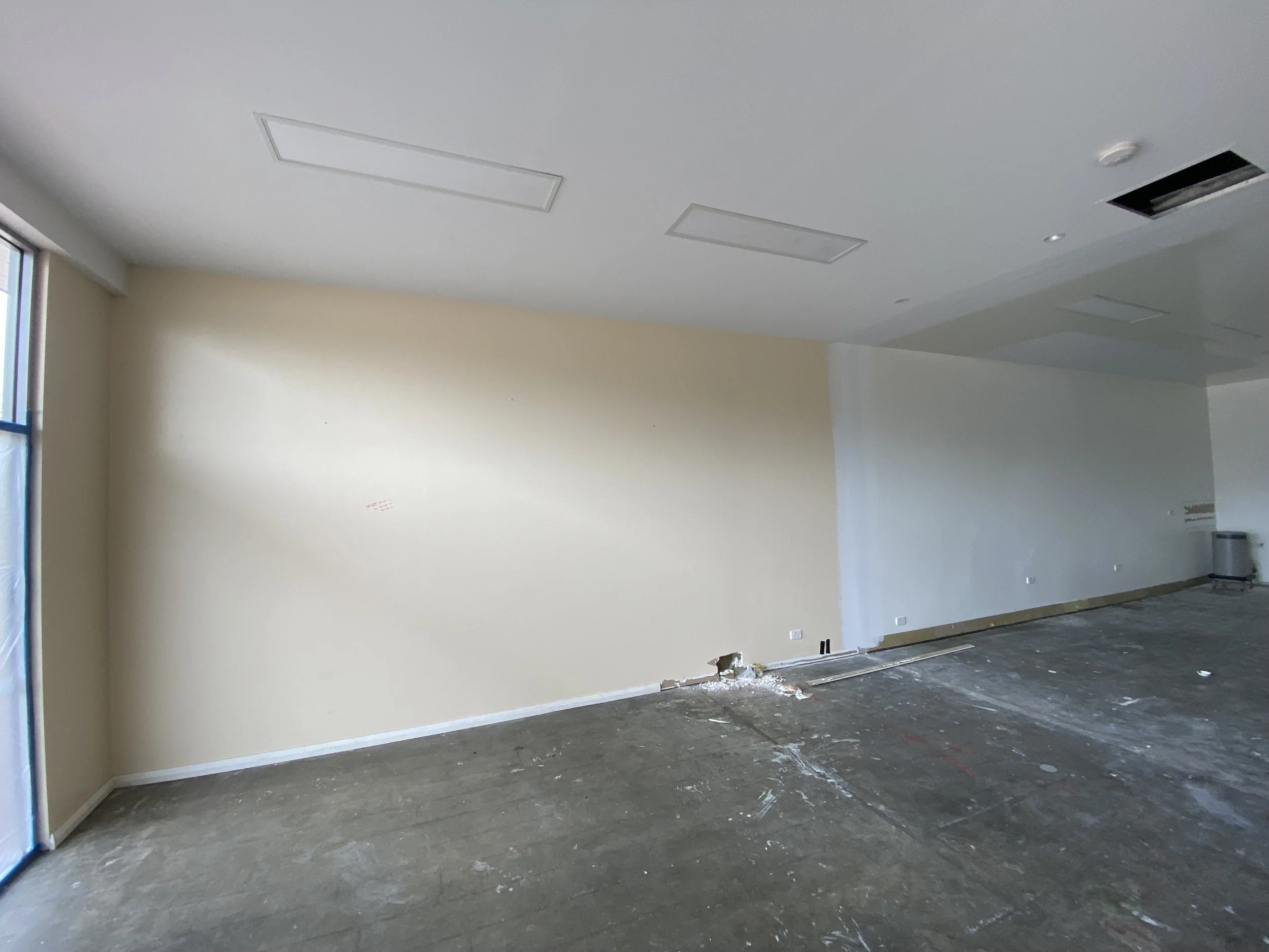 Empty room under construction or renovation with unfinished flooring, a white wall, and partially installed ceiling fixtures.