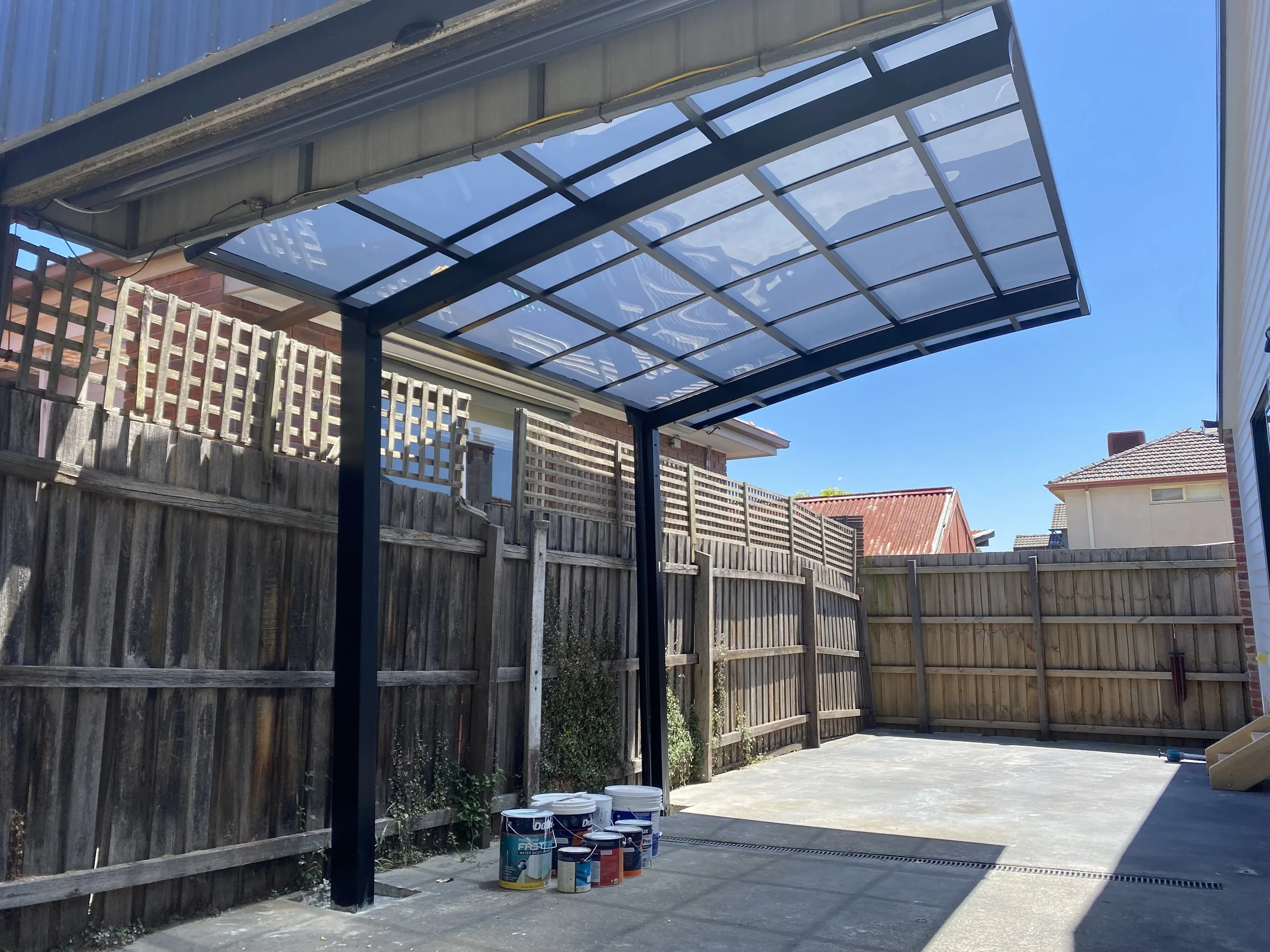 A backyard with a wooden fence, a concrete patio, and a blue metal and glass awning structure. Several paint cans are on the ground, and neighboring houses are visible under a clear blue sky.