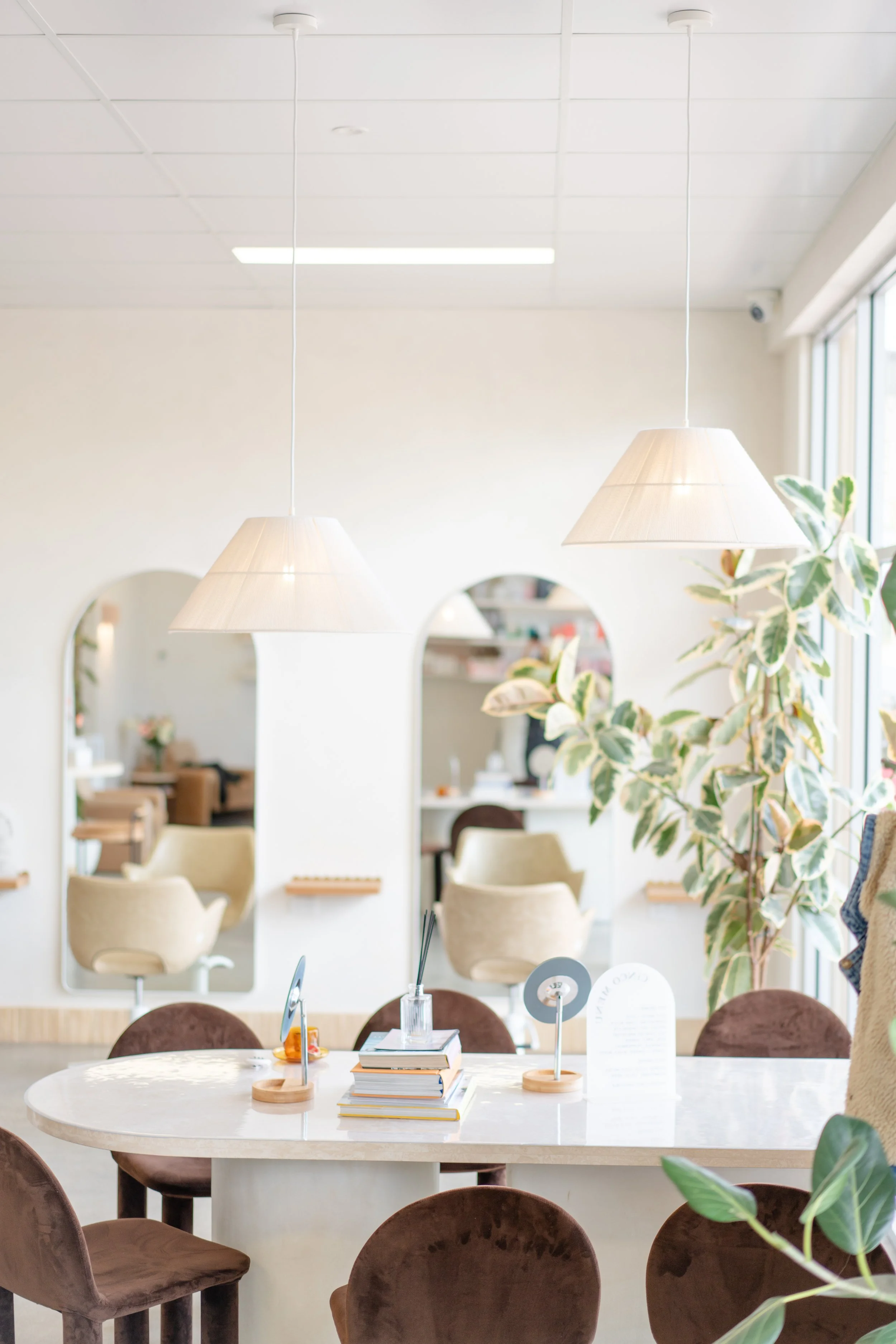 A bright, modern dining area with a round white table surrounded by brown velvet chairs. Shelves and mirrors with rounded edges and a large window with vertical blinds are visible in the background. Potted plants are near the window, adding greenery 