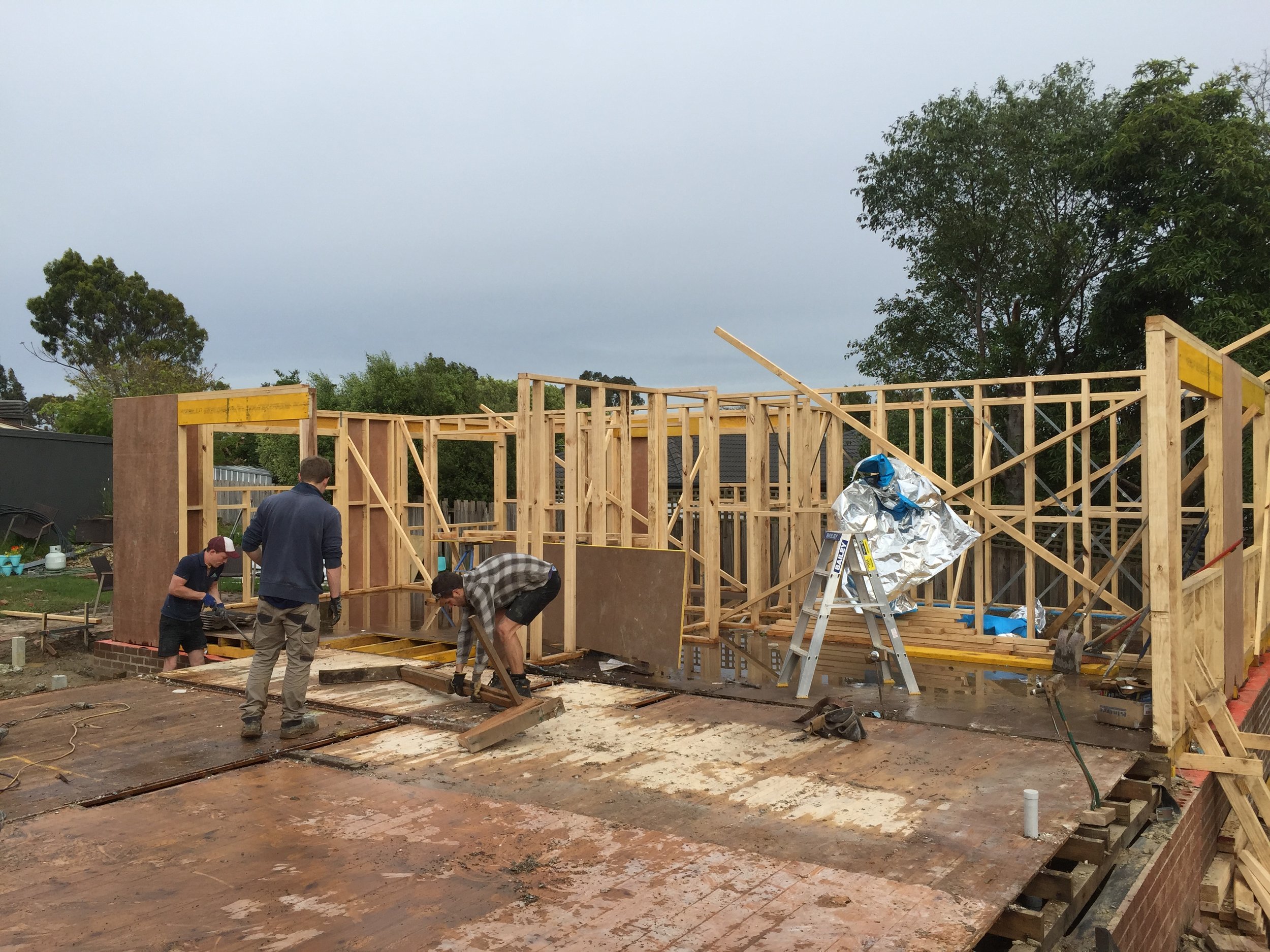 Construction workers building the wooden framework of a house in a backyard neighborhood. The sky is overcast, and there are trees and some rooftops in the background.