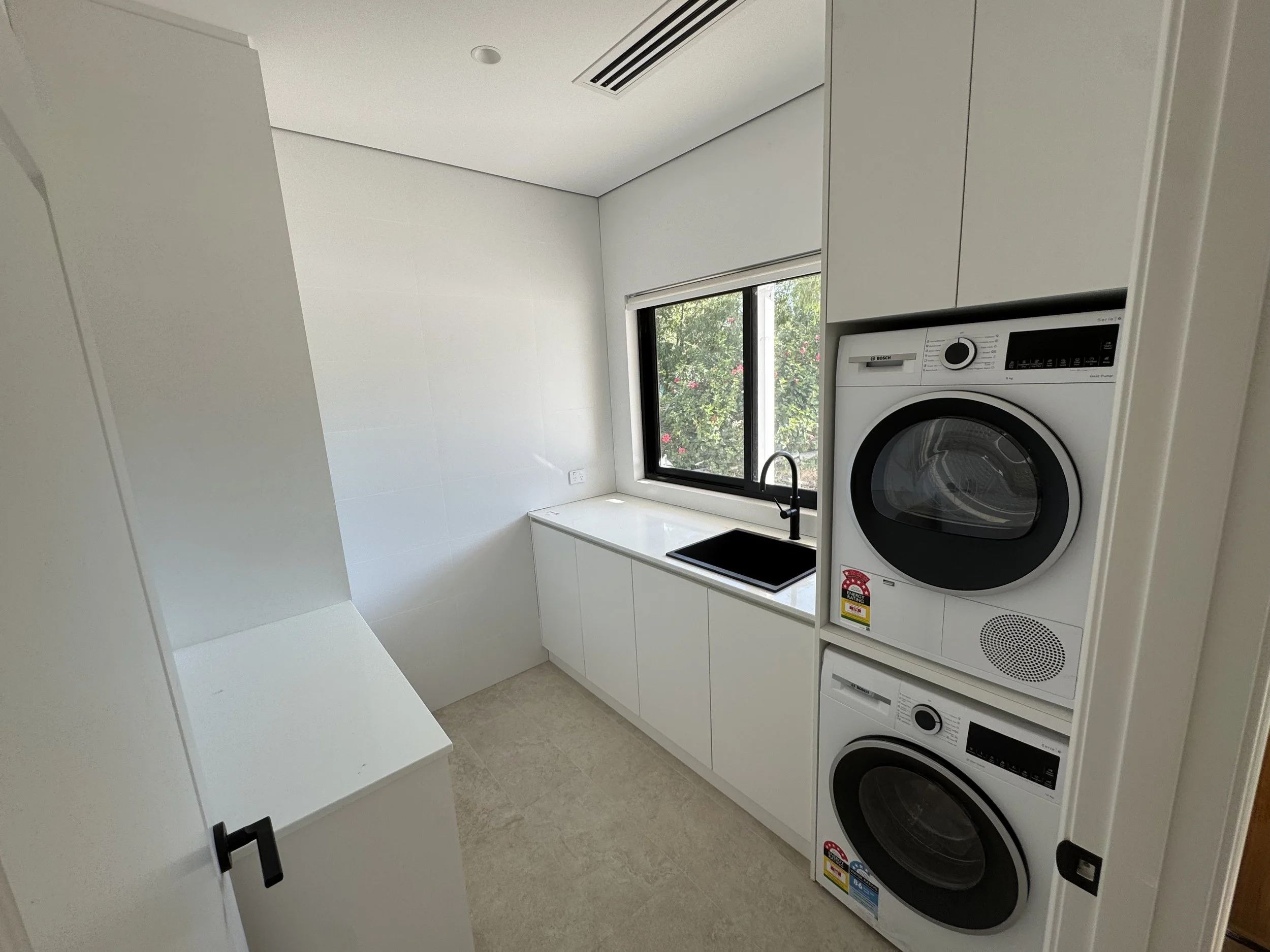 A minimalist laundry room with white walls, a white countertop, a window with views of greenery, a black sink faucet, and a stack of two Bosch front-loading washing machines and dryers.