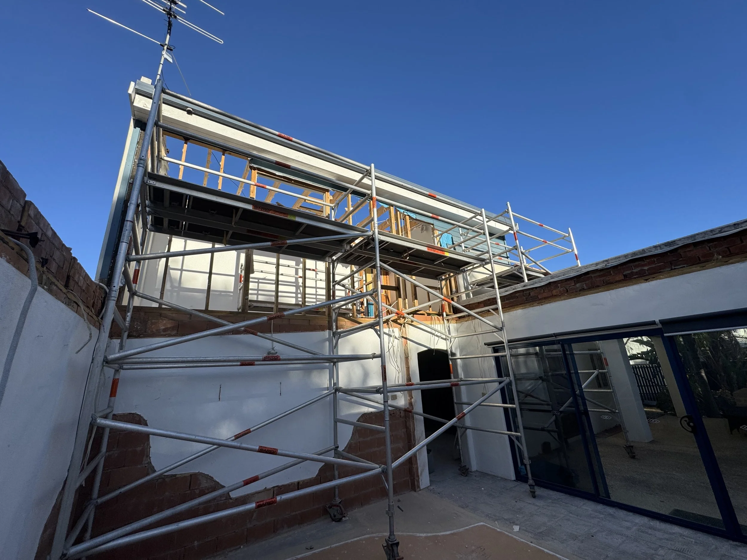 Construction scaffolding around a building with exposed brick and a large window frame, under a clear blue sky.