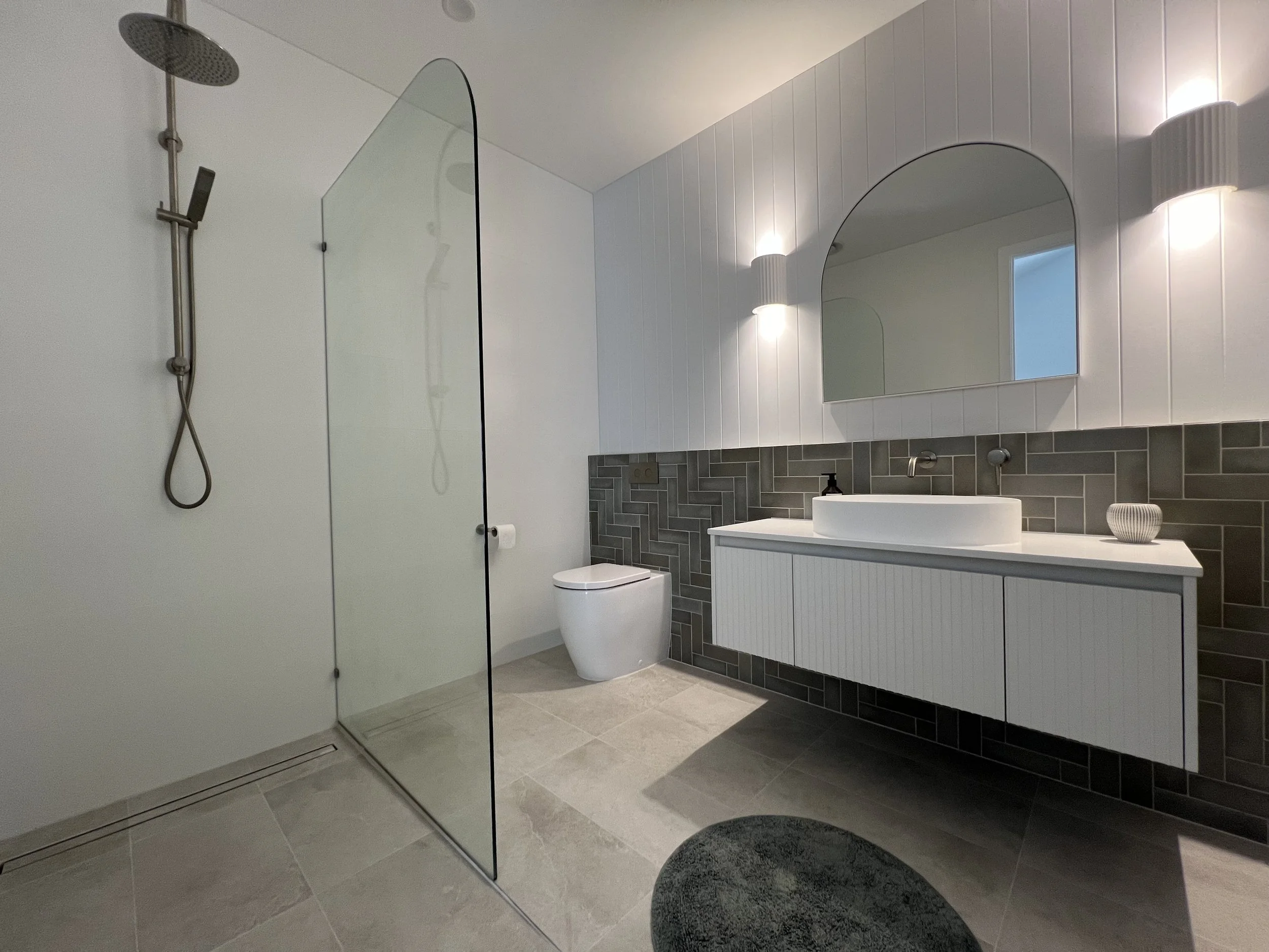 A modern bathroom with a walk-in shower, a white toilet, a white vanity with a vessel sink, and a large mirror. The walls are white with vertical paneling, and there are two wall-mounted lights.