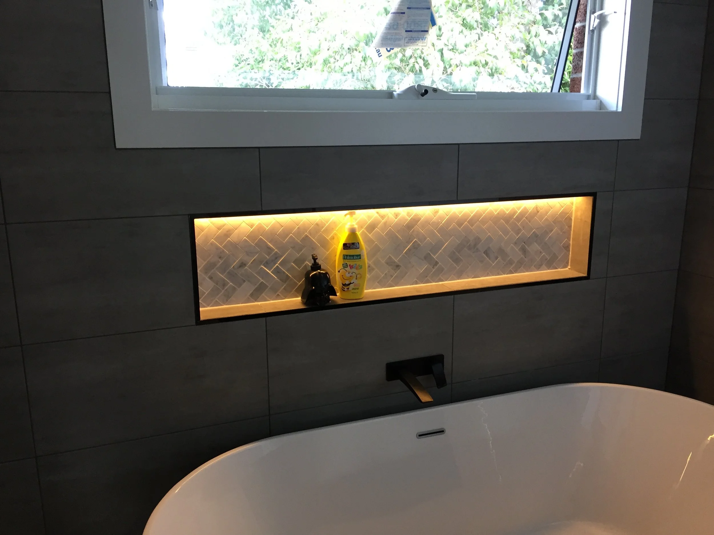A bathroom with a large window showing greenery outside, a built-in shelf with a yellow bottle of cleaning product, a faucet, and a view of the bathtub in the foreground.