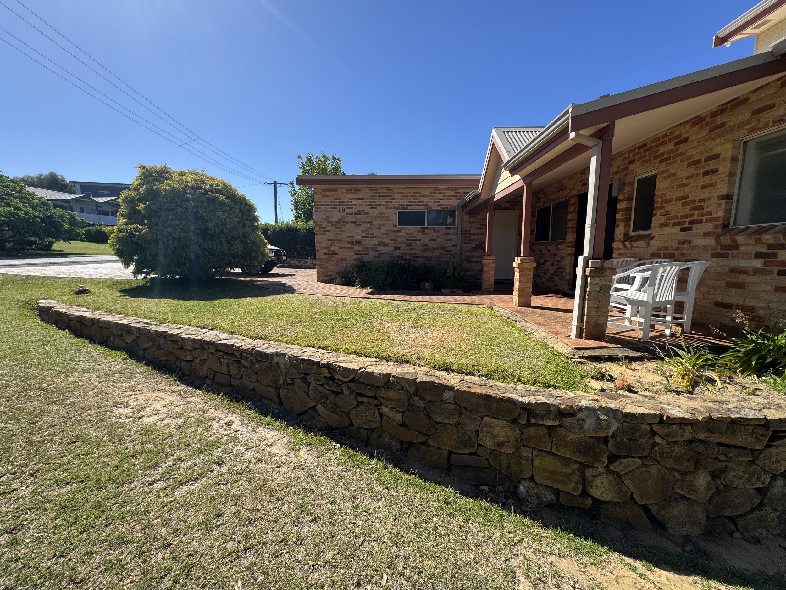 A brick house with a porch, white chairs, and a brick driveway. A large bush and grass area in the front yard. Power lines and a neighboring house visible in the background under a clear, sunny sky.