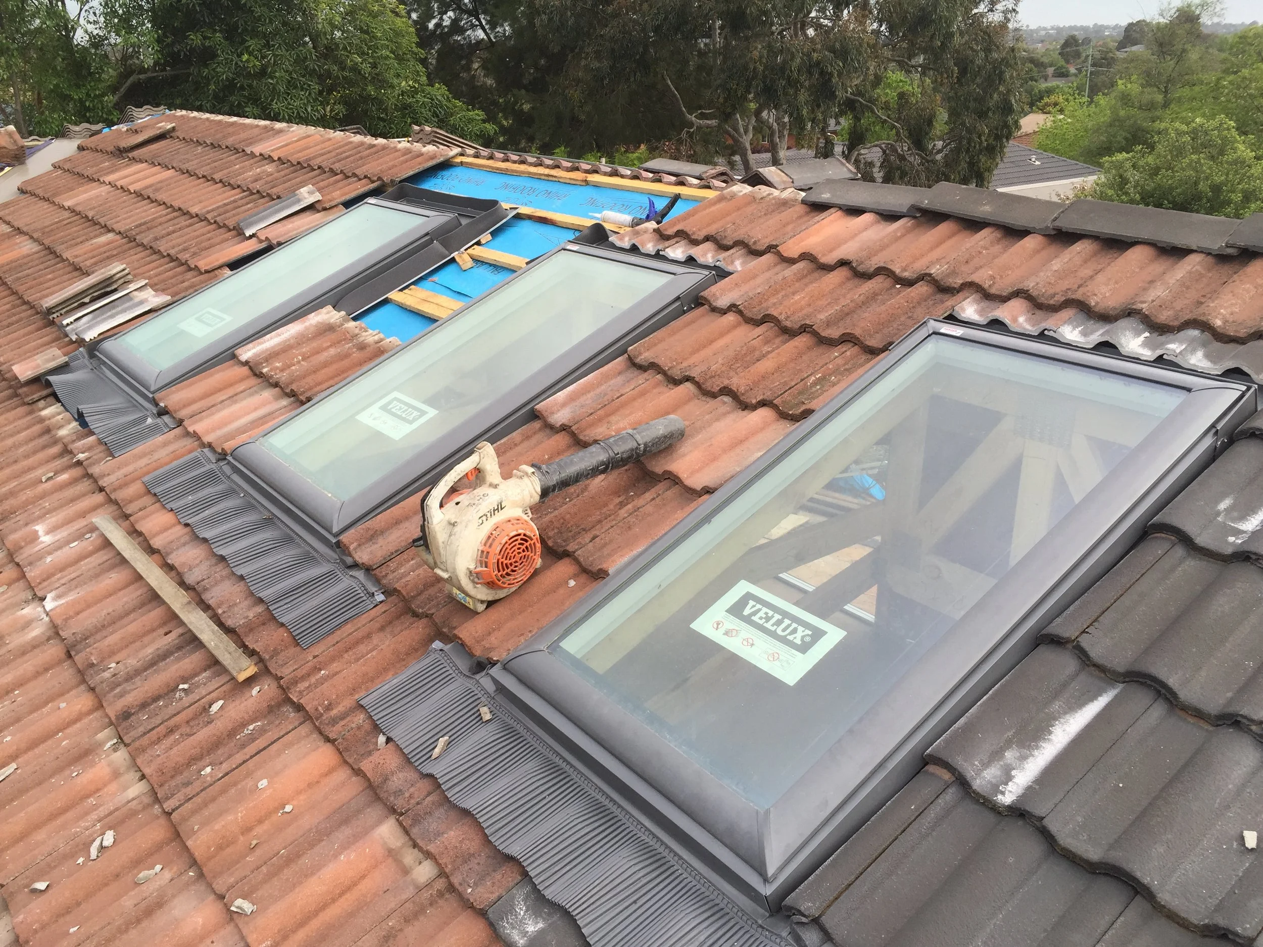 A roof under construction or repair with old and new roofing materials. Several skylights are installed among the tiles. In the background are trees and neighboring houses.