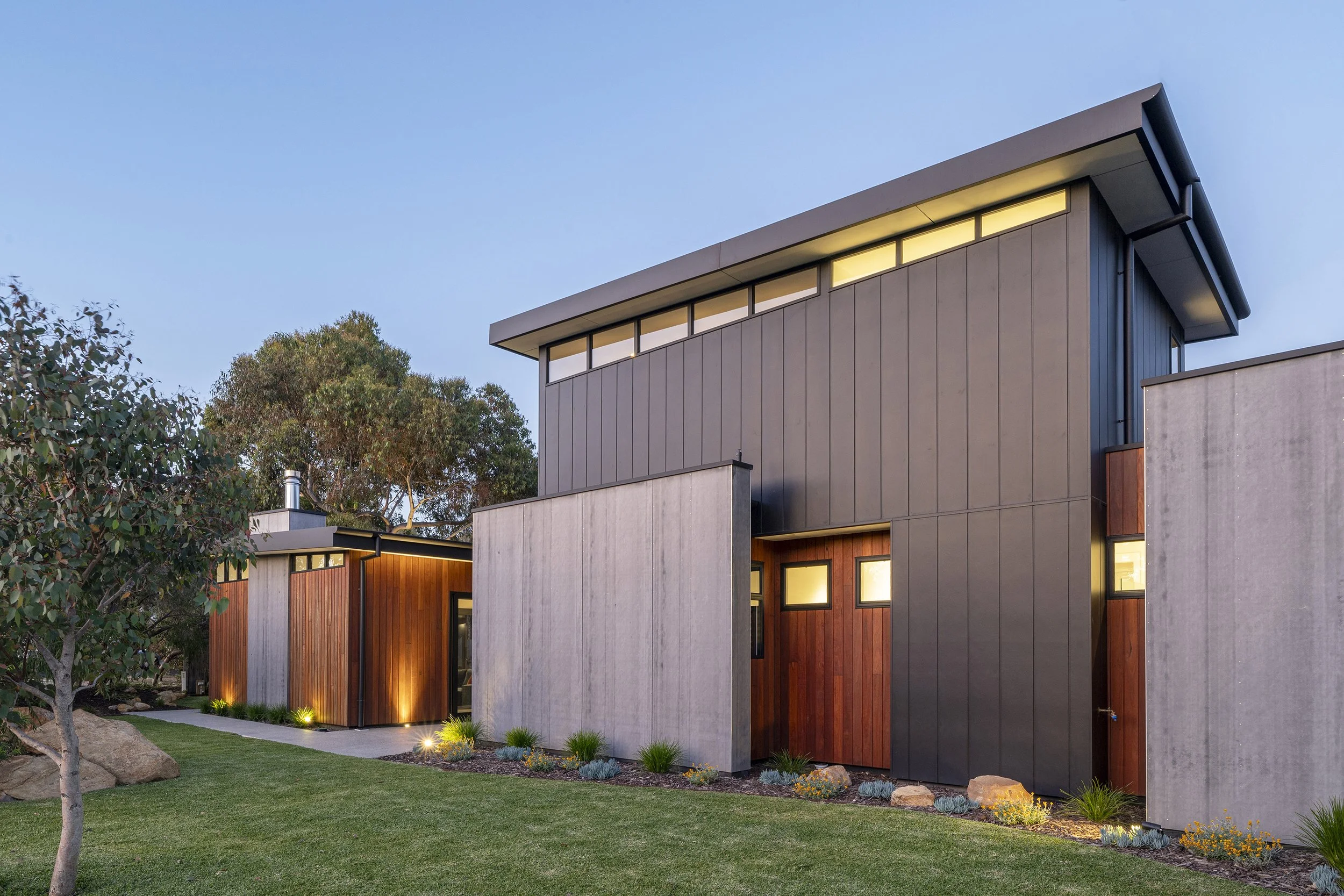 Modern multi-story house with large windows, wood and concrete exterior, surrounded by landscaped yard with grass, small plants, and trees.