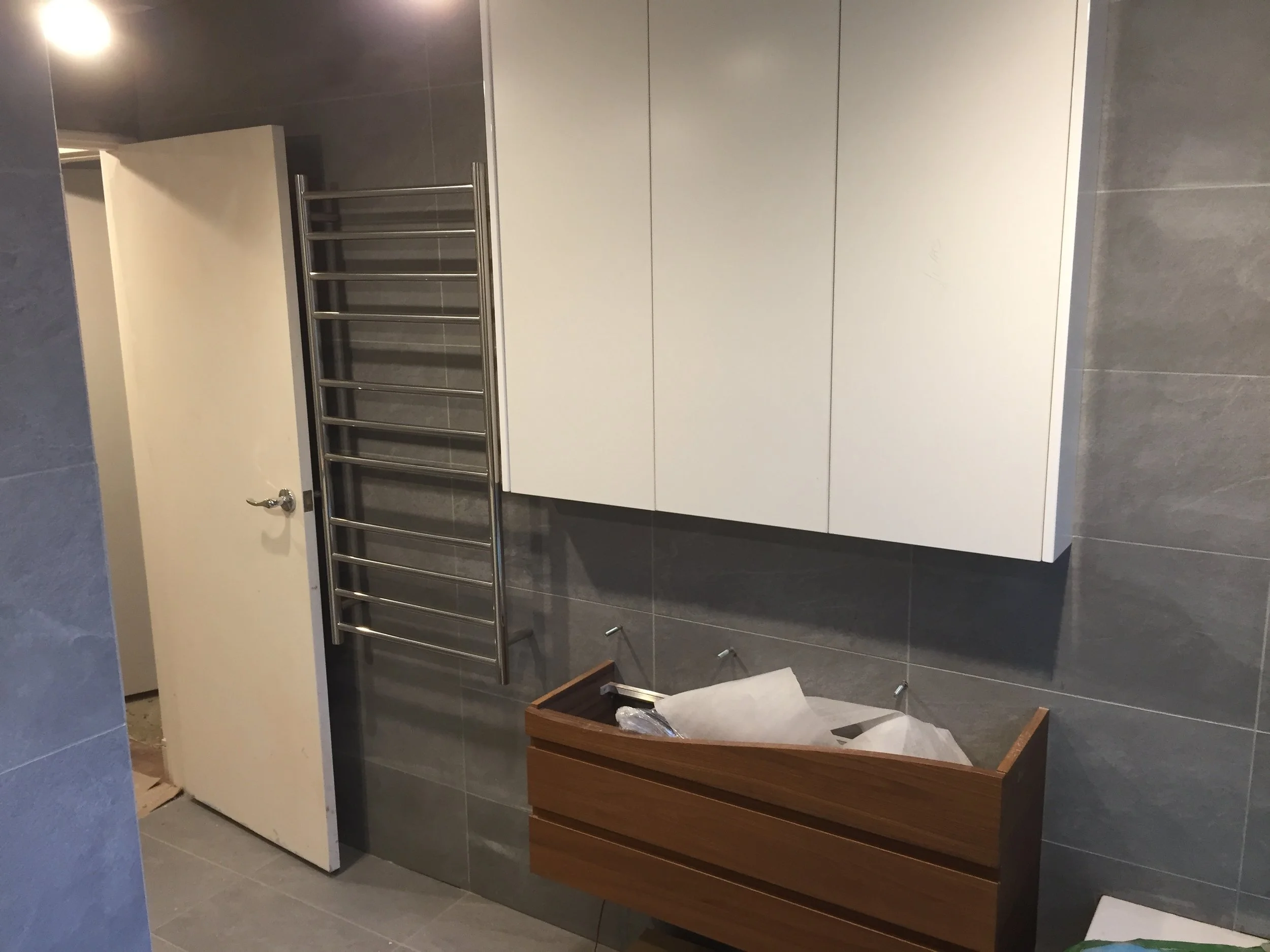 Bathroom with gray tiled walls and floor, white cabinet with several doors, a towel radiator, a small wooden cabinet, and a partially open door.