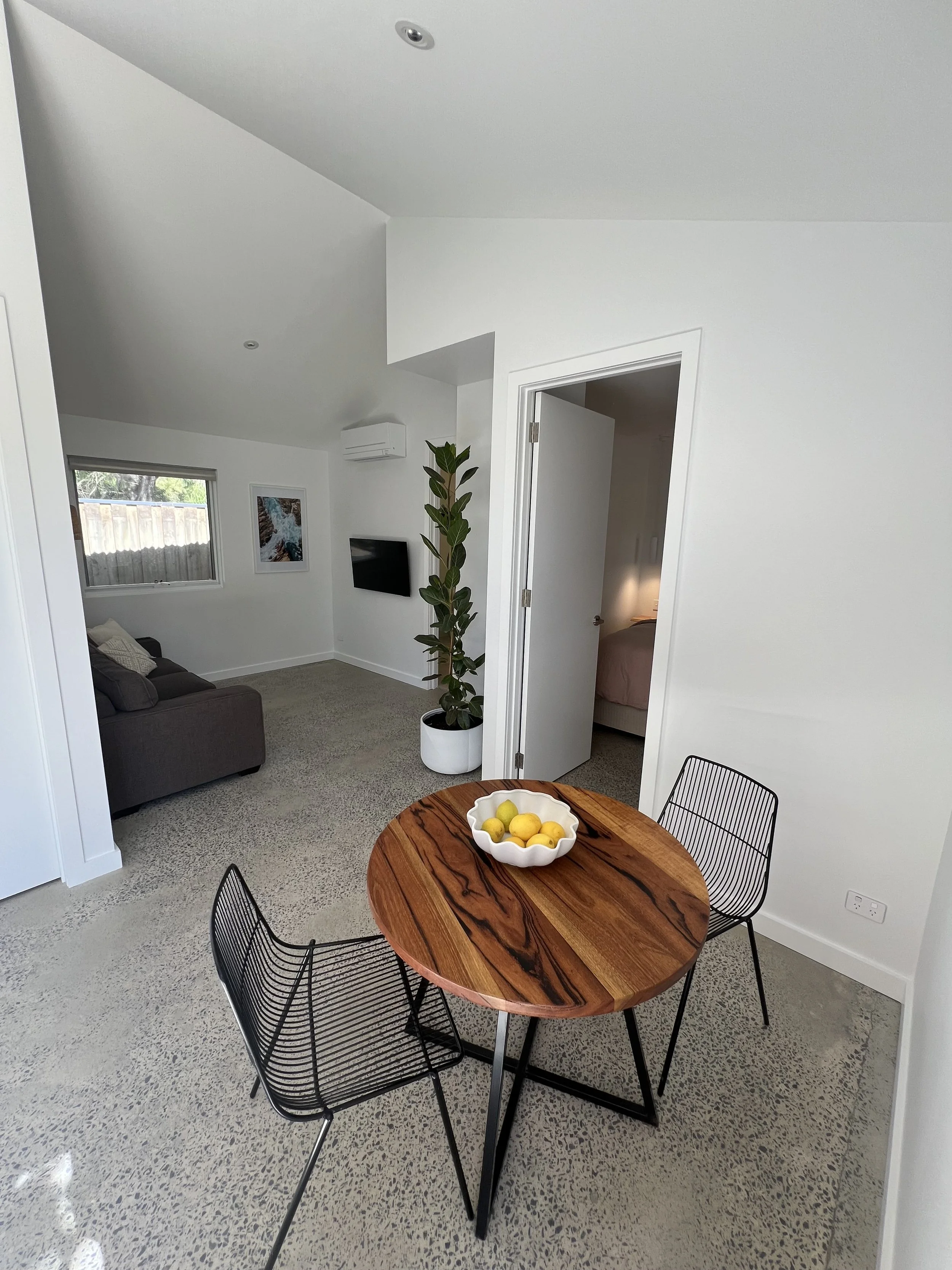 A round wooden dining table with three black wire chairs, a white bowl with lemons and limes, in a modern living room with a gray couch, a window, a framed picture, a television, a potted plant, and an open door leading to a bedroom.