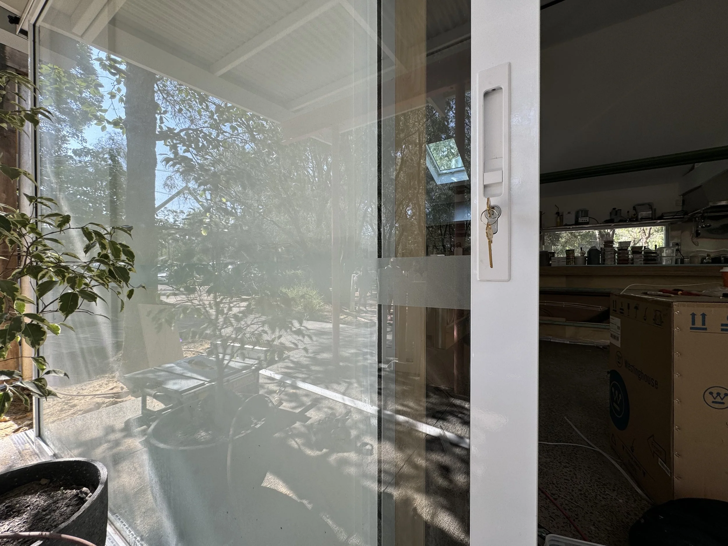 View of a house interior with a large glass window reflecting trees outside, flowering plant in the foreground, partially open white door with a key in lock, and a glimpse of a kitchen area with cabinets and counters.