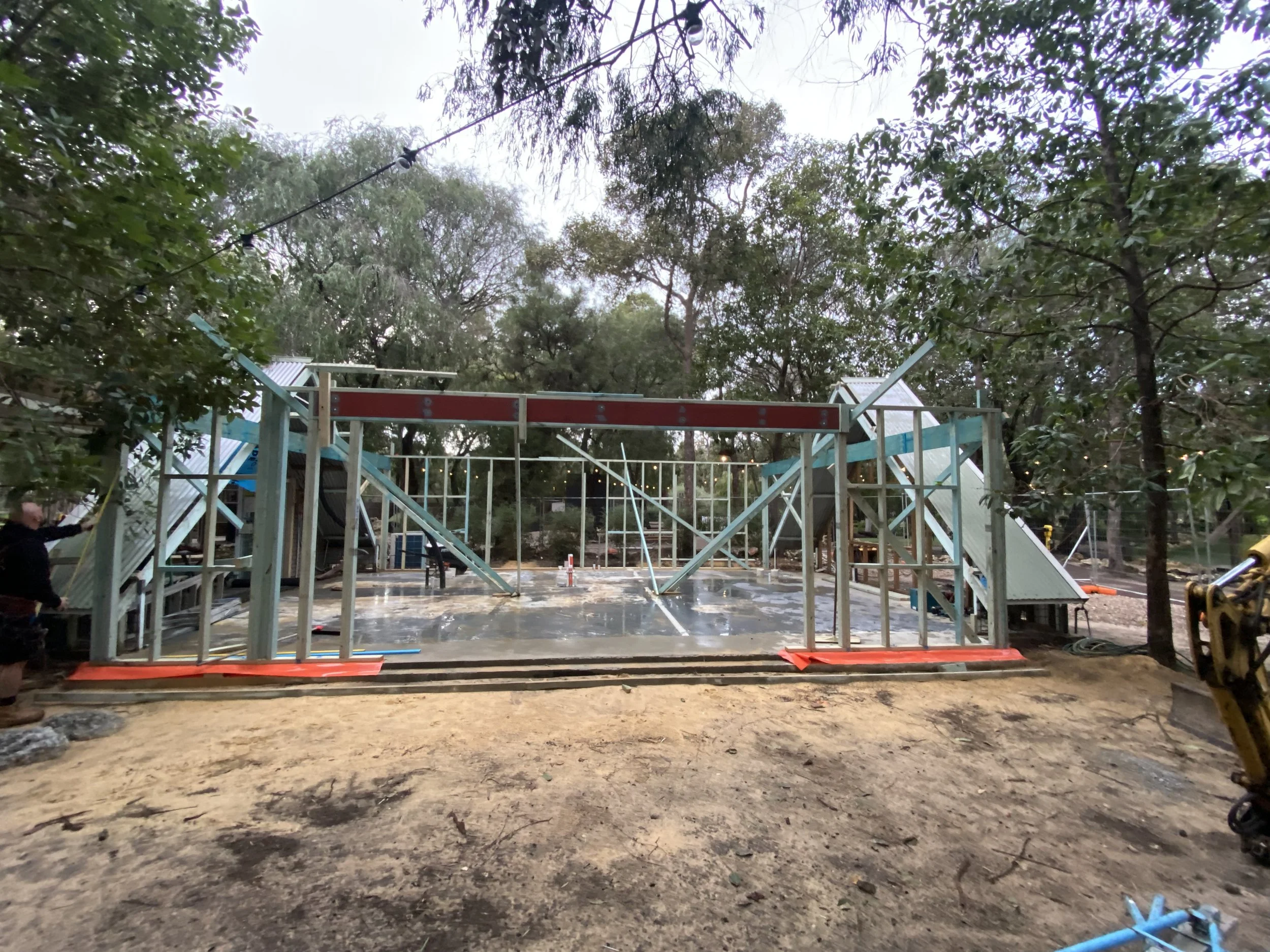 Under construction outdoor structure with framing in progress, surrounded by trees, with a person on the left holding an object, on a dirt ground.