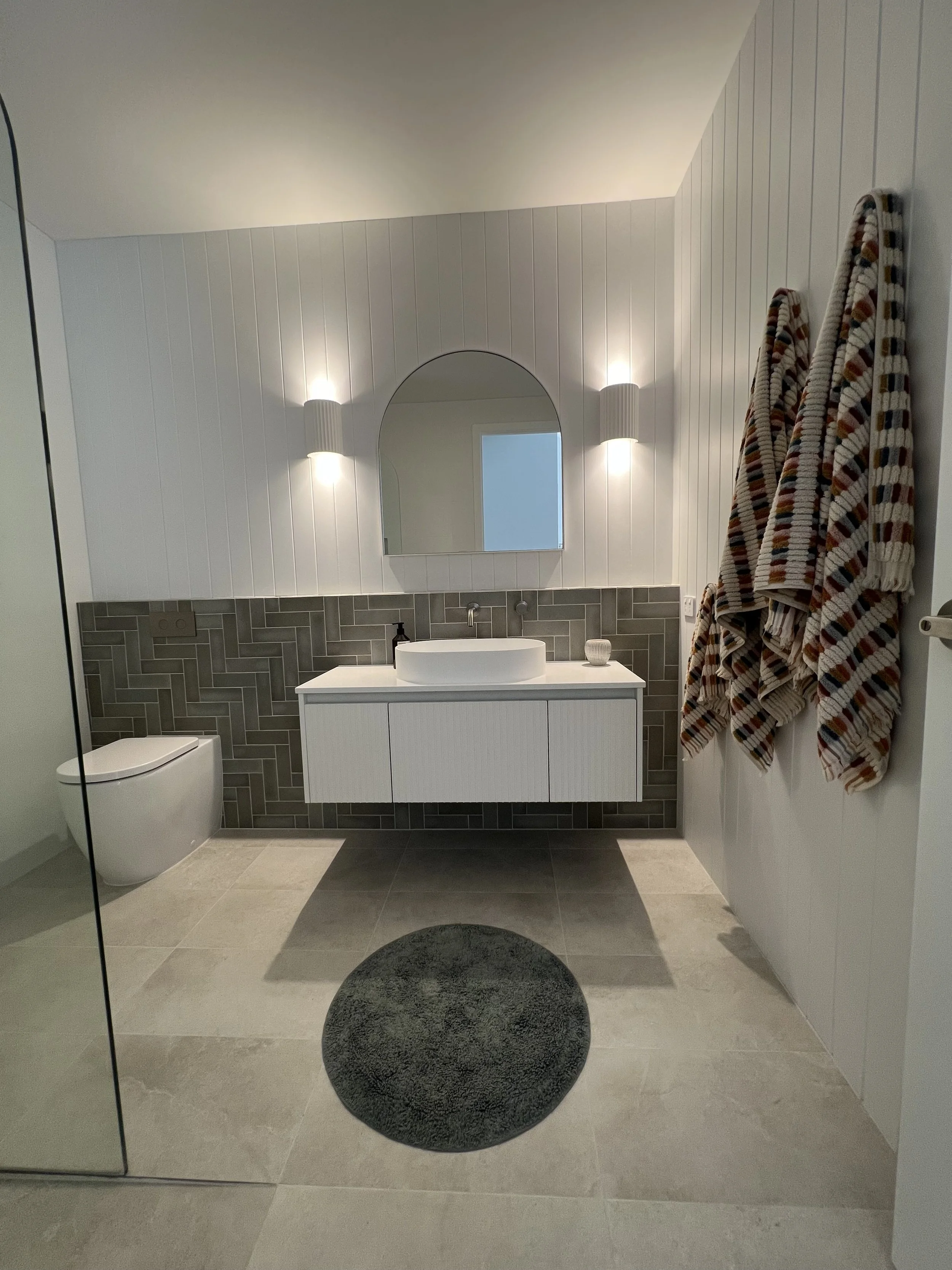 Modern bathroom with a white vanity, a round vessel sink, gray tiled backsplash, mirror, wall-mounted lights, towels hanging on the side, and a dark round rug on gray floor tiles.