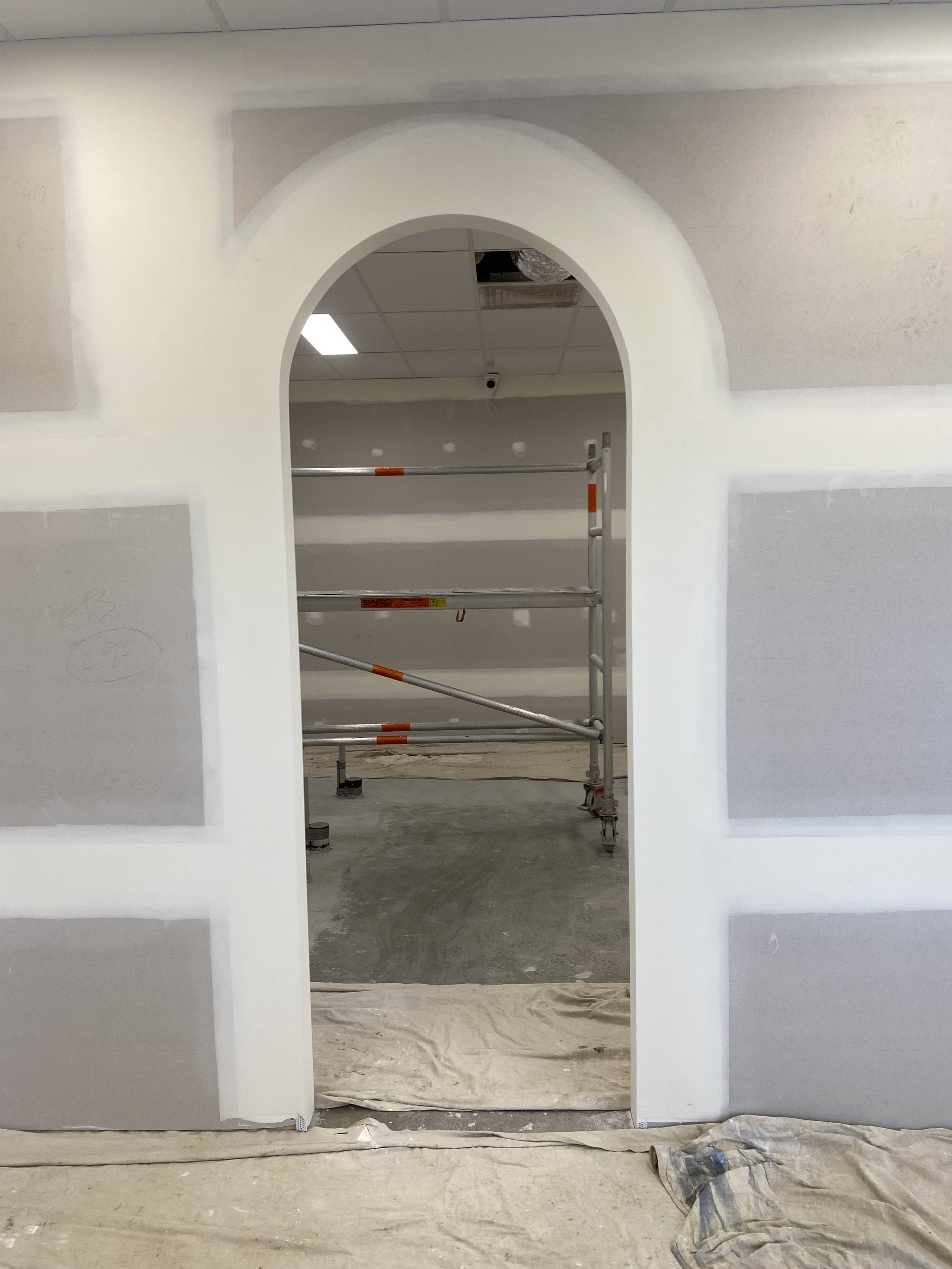 Interior of a building under construction with an arched doorway, scaffolding, and unpainted drywall with seams visible, and protective covering on the floor.