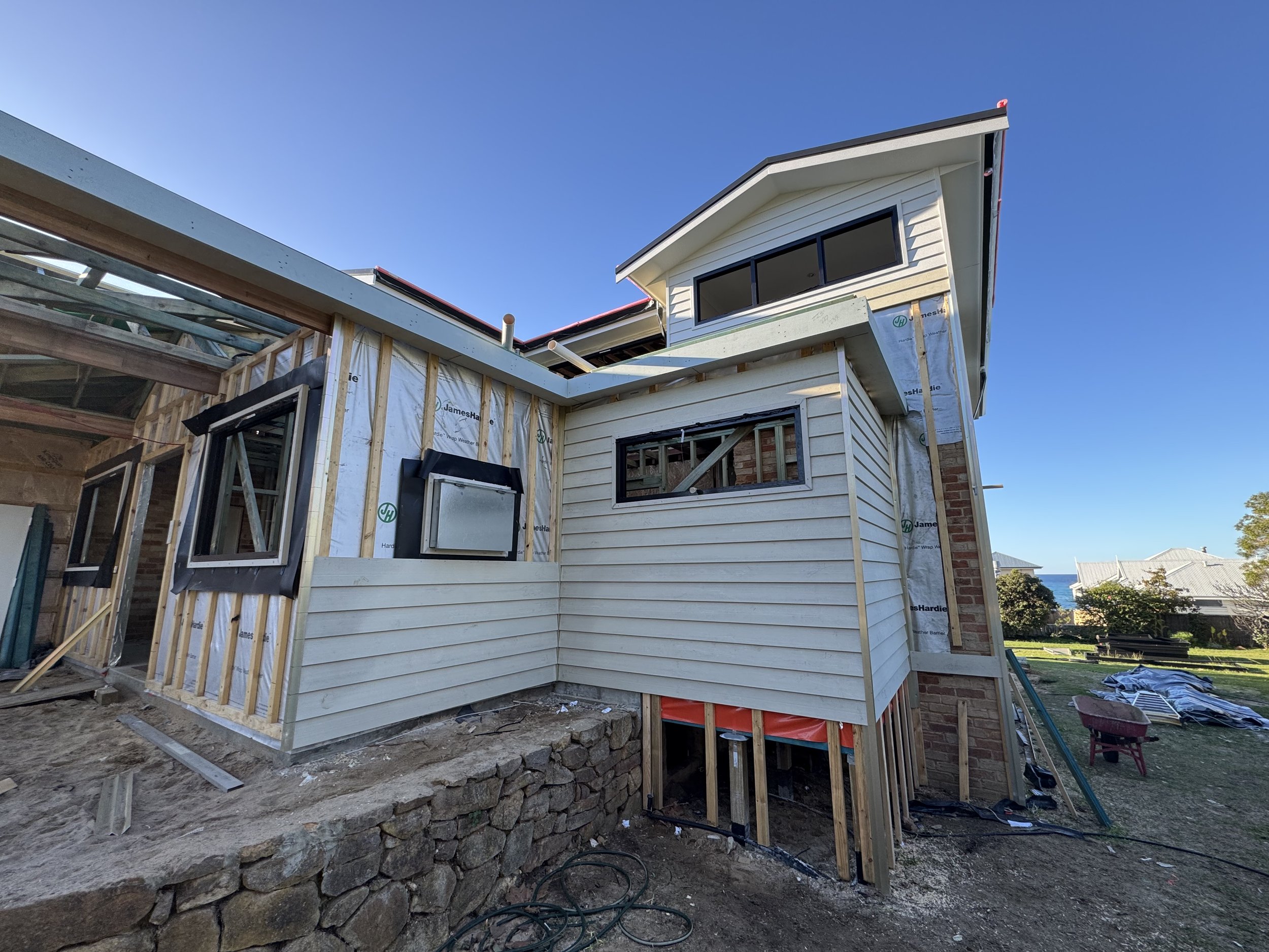 Under construction house with wooden framing, a second-story window, and siding being installed, against a clear blue sky.