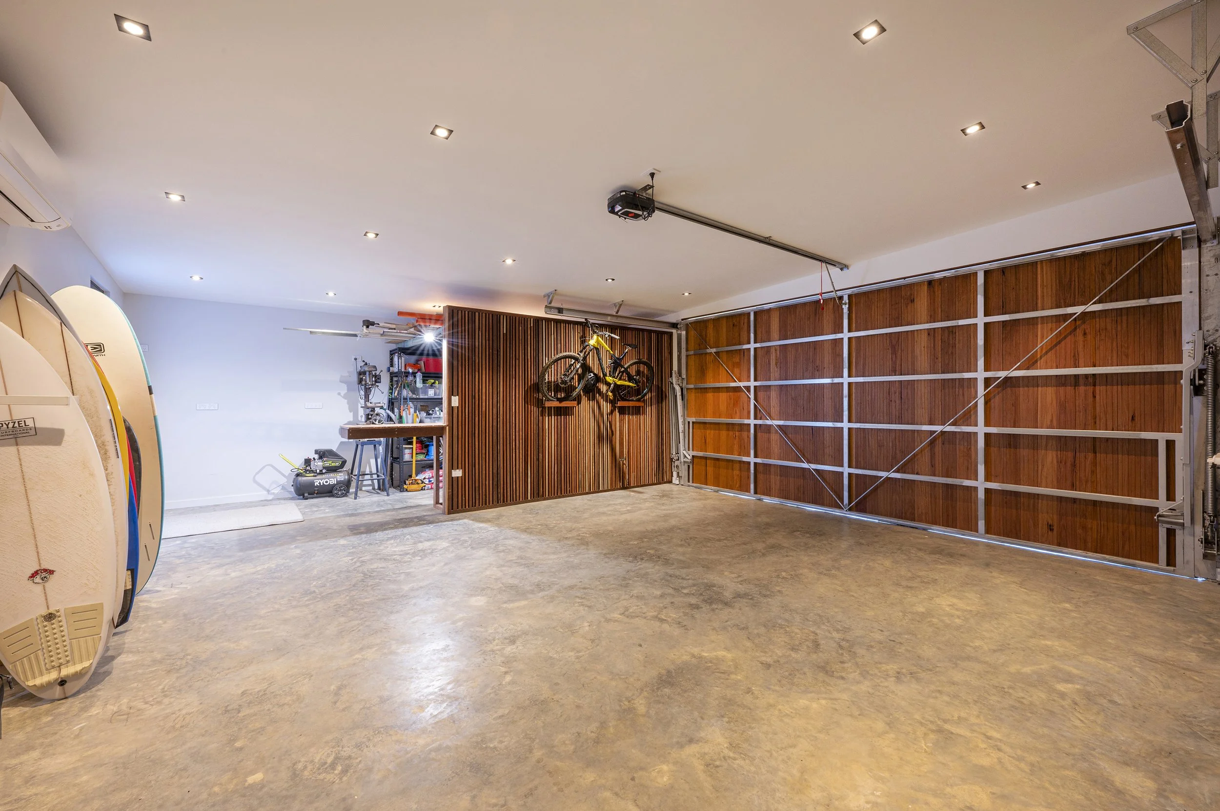 A spacious garage with a wooden sectional garage door, several paddleboards leaning against the wall, and a bicycle mounted on a wall rack. The garage has a polished concrete floor and recessed ceiling lights.